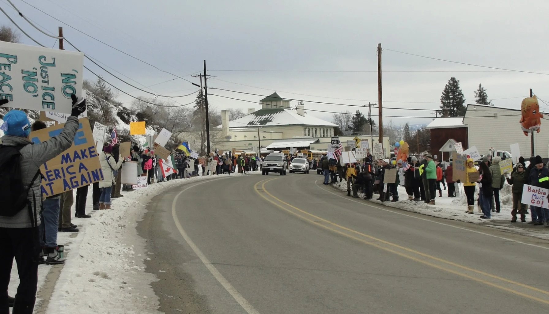 Twisp “Stand With Us" protesting along a snowy street, holding signs and balloons, with some signs reading 'NO JUSTICE NO PEACE' and 'MAKE AMERICA GREAT AGAIN' amid a snowy landscape and rural buildings.