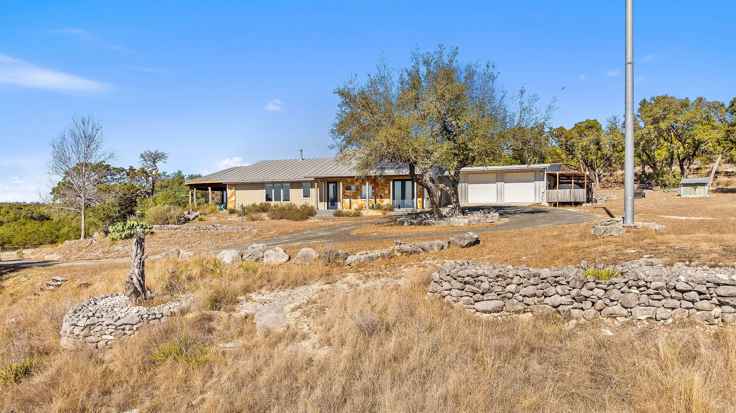 A rustic-modern home in the Texas Hill Country with a tree in the front yard and a stone retaining wall. A gravel path leads to the house, and there is a large tree to the right of the house with a garage attached. The sky is blue with a few clouds.