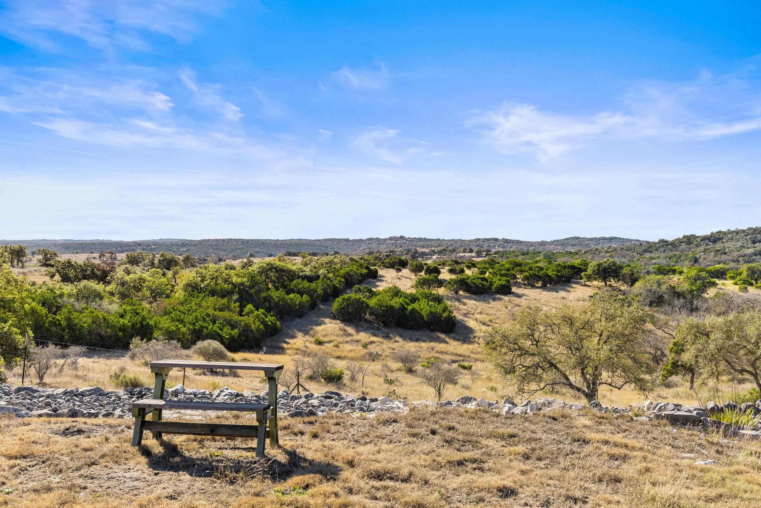 A bench in a dry grassy field overlooking rolling hills within the Texas Hill Country covered with green shrubs and trees, under a partly cloudy blue sky.