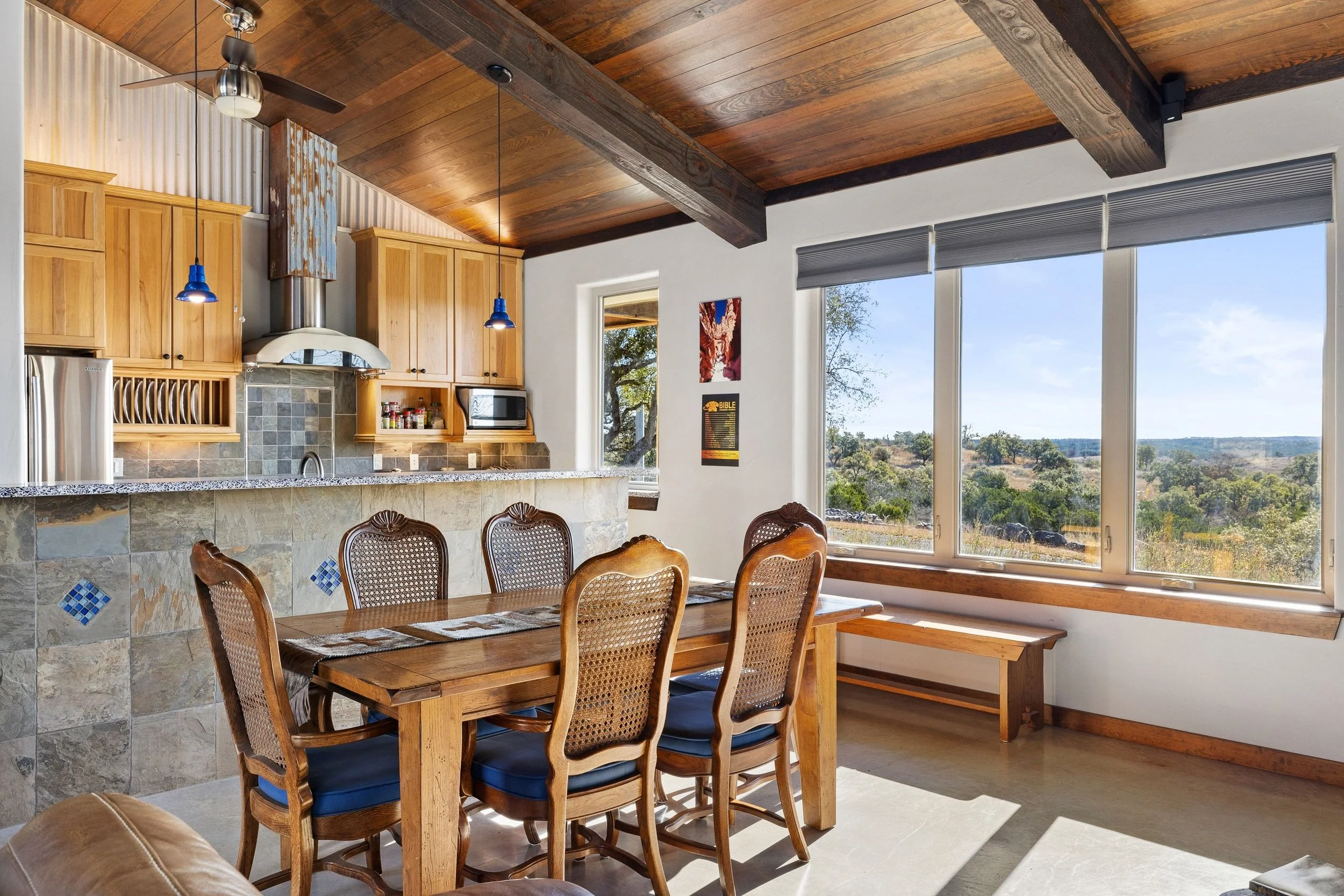 Dining area with a wooden table and six chairs, large window with a scenic view, kitchen with wooden cabinets and a breakfast bar, and a wooden ceiling with exposed beams.