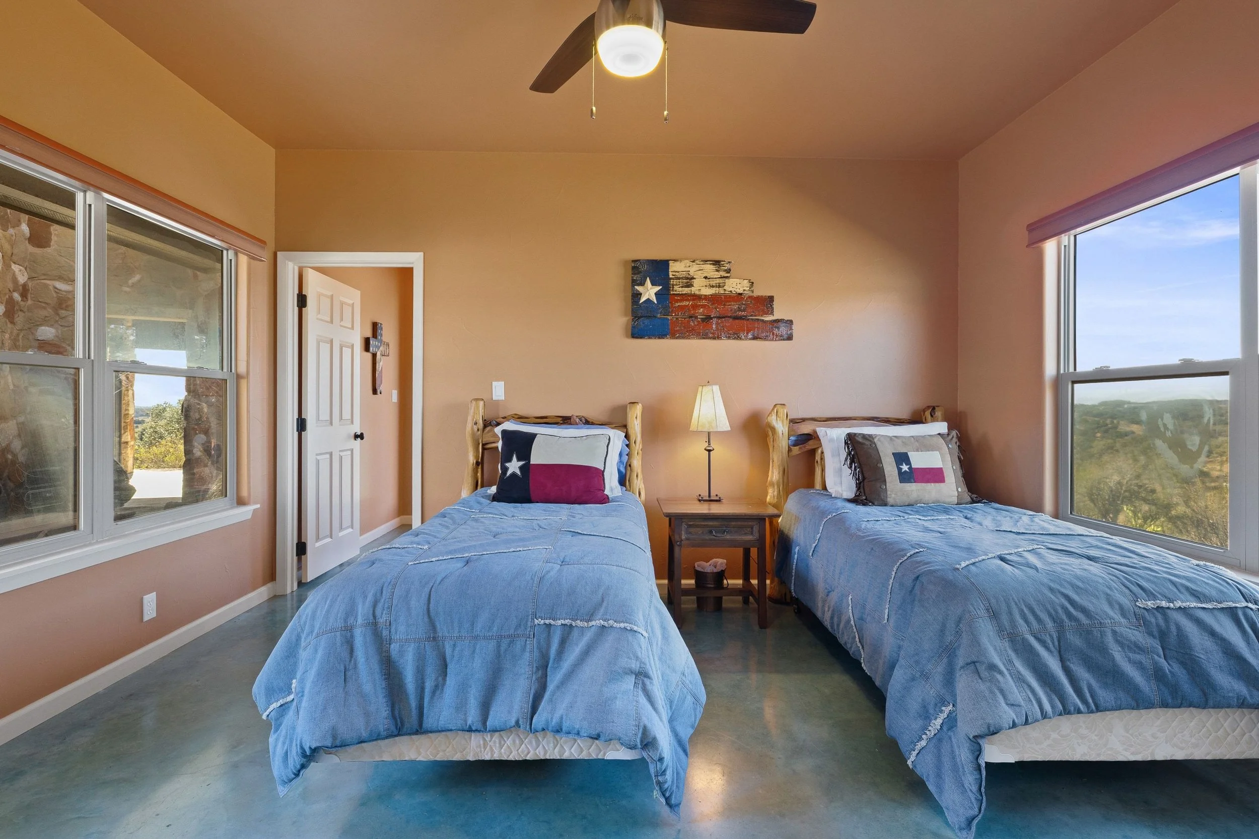 A bedroom with two twin beds, each with Texas flag-themed pillows, a small wooden nightstand between them with a lamp, a Texas flag artwork on the wall, beige walls, large windows showing outdoor greenery, ceiling fan, polished concrete floor.