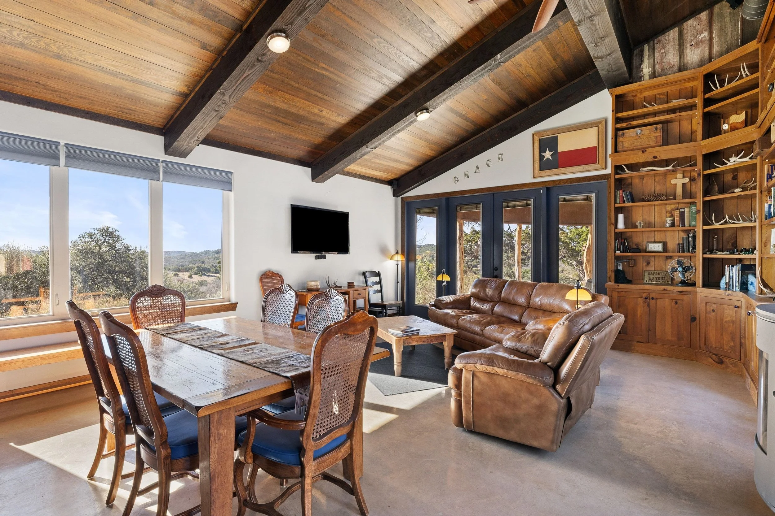 Living room featuring a dining table with six chairs, a leather sofa, a wooden coffee table, a wall-mounted TV, large windows showcasing an outdoor landscape, and built-in wooden shelves with decor and books, with a wood-paneled ceiling.