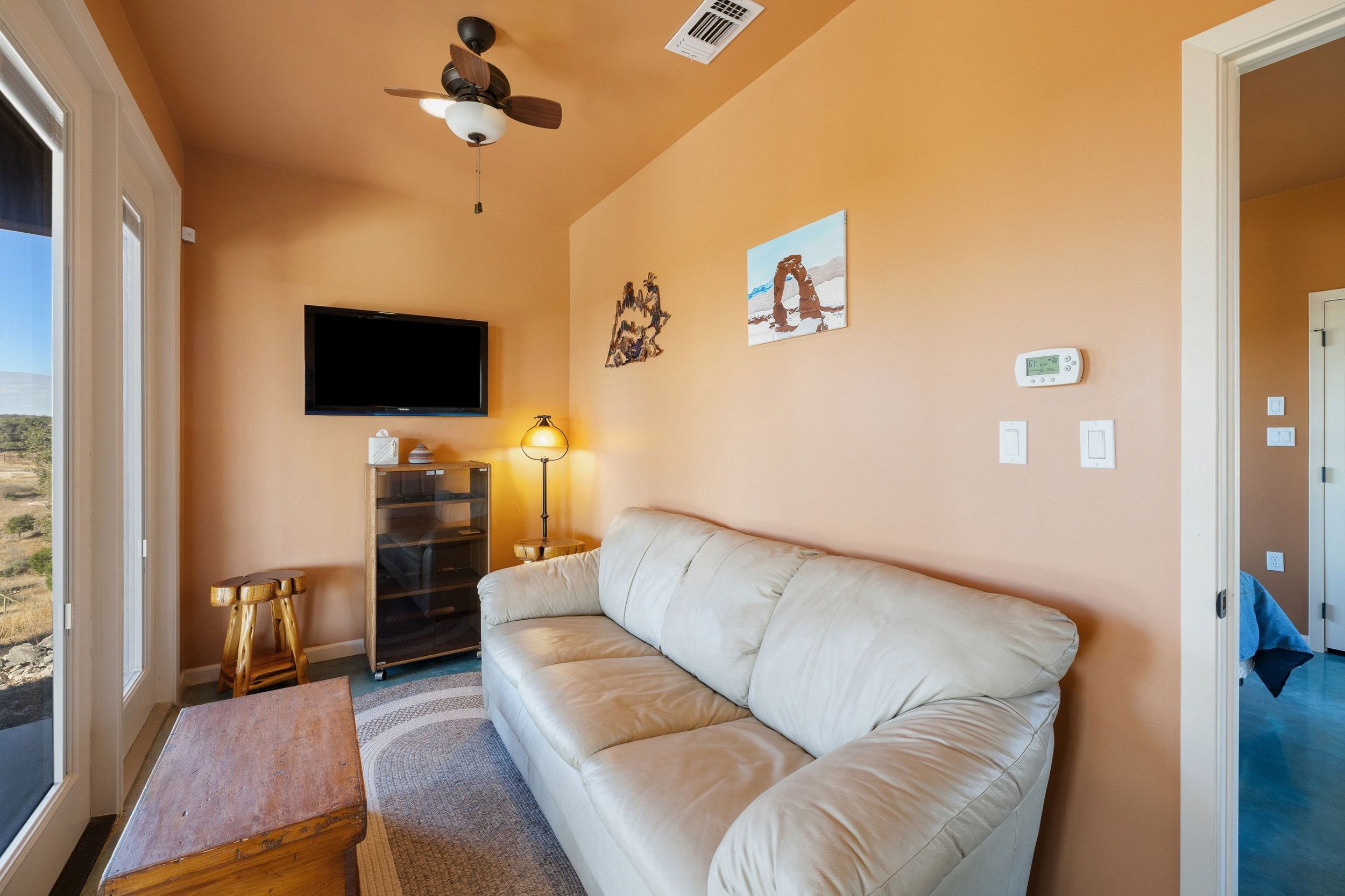 Living room with beige couch, wall art, ceiling fan, TV, standing lamp, and wooden furniture, with a view outside through glass door.