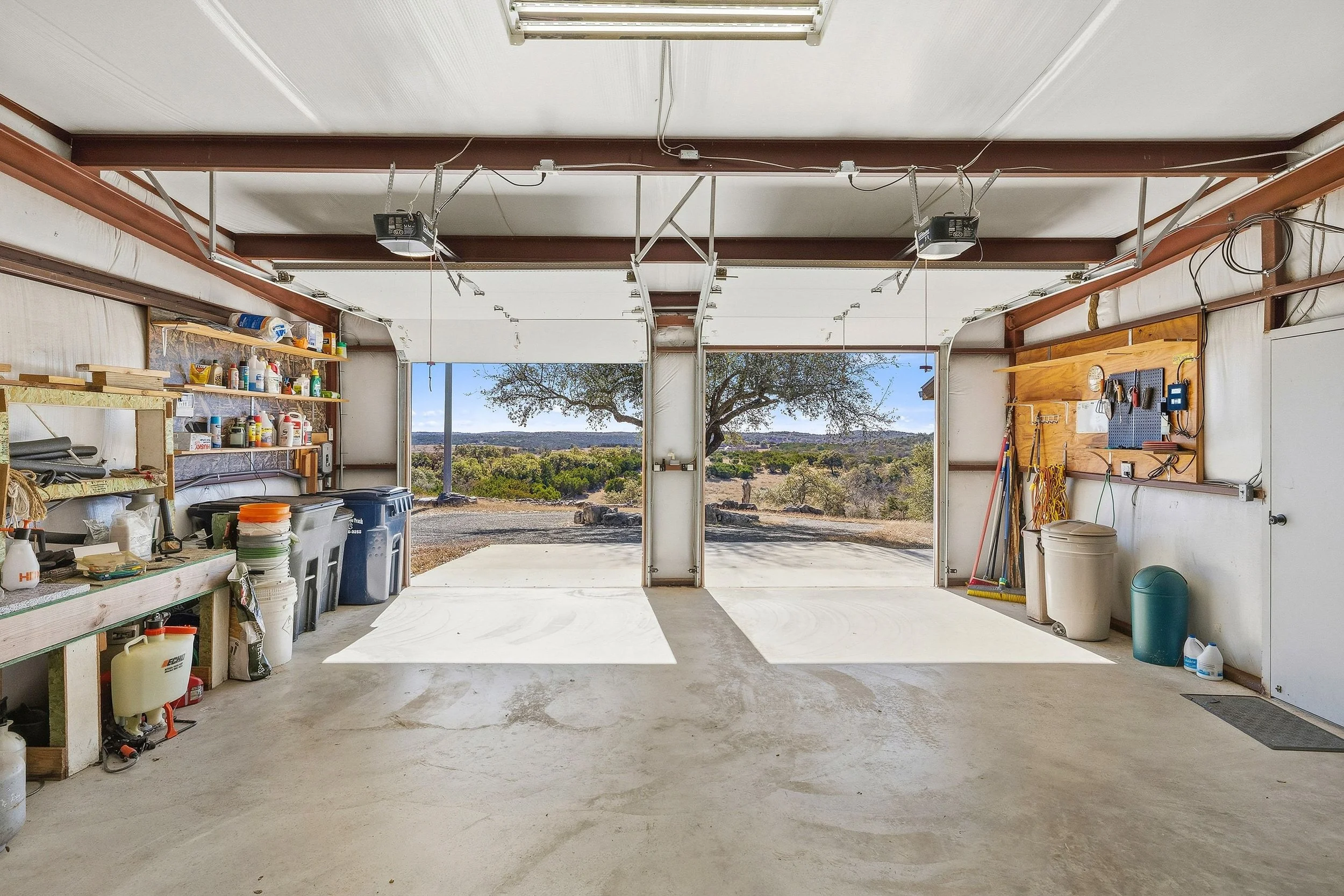 A spacious garage with an open door revealing a view of trees and Hill Country landscape outside.