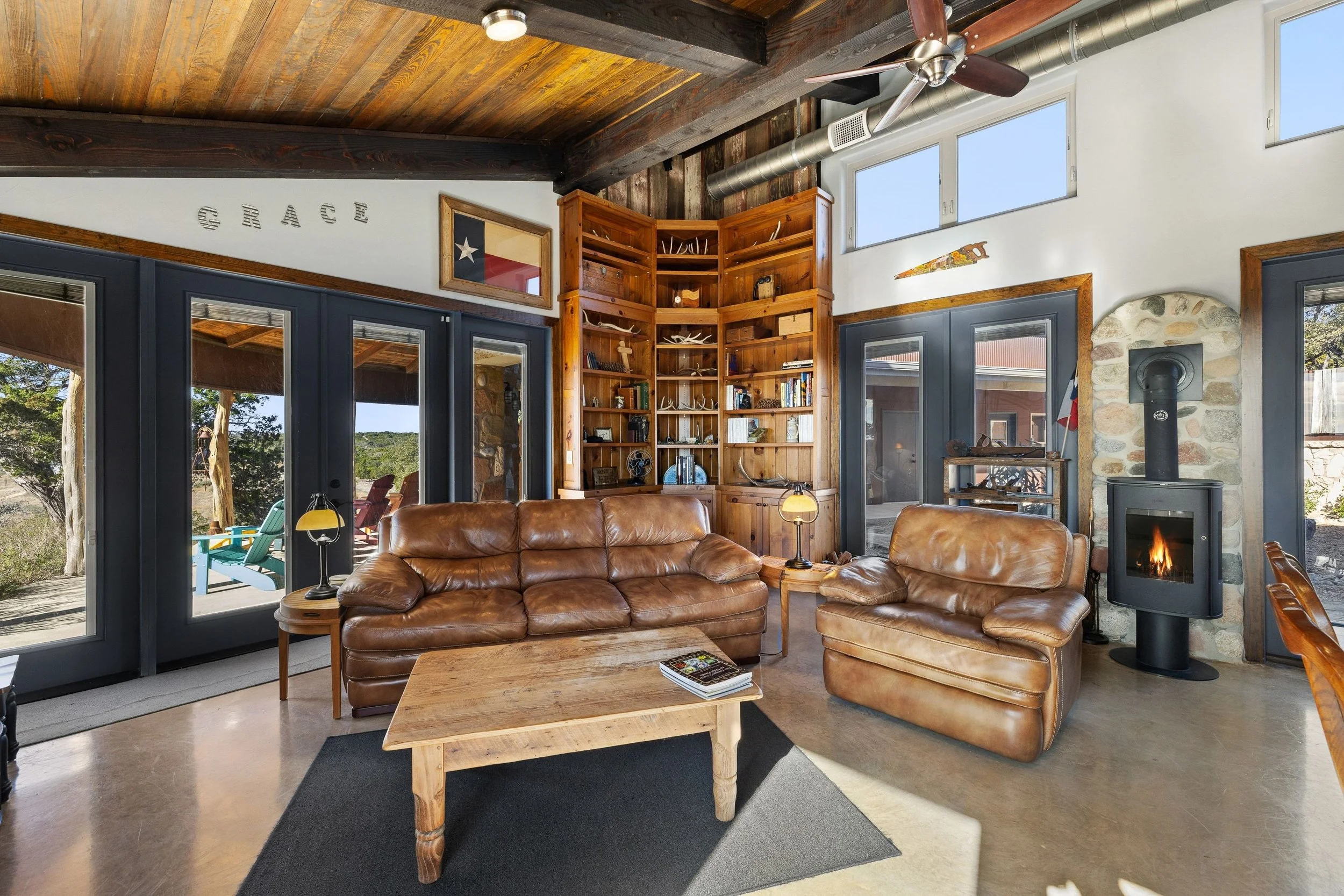 Living room with leather sofas, a wood coffee table, built-in wooden shelves, a stone fireplace with a wood stove, ceiling fan, and large windows showing outdoor scenery.