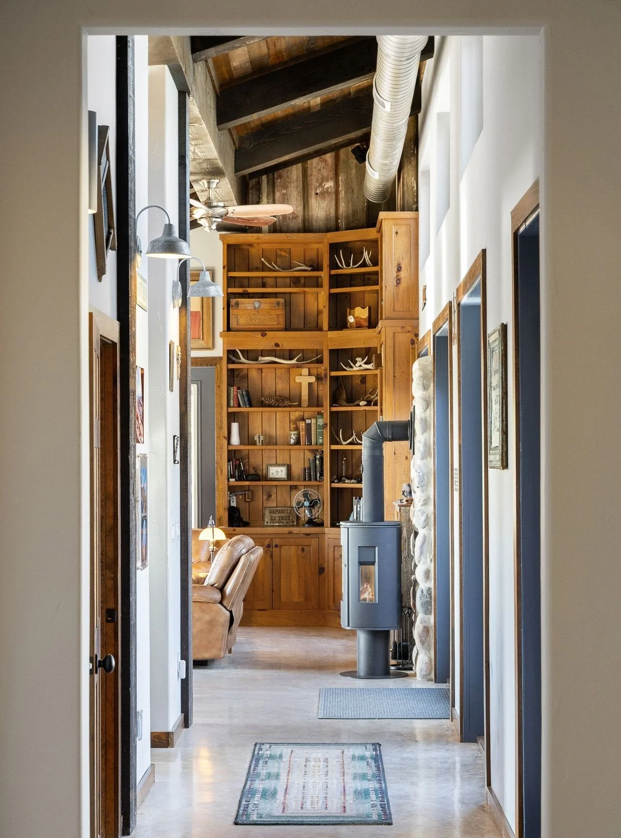View through a doorway into a cozy living room with wooden shelves, antlers, books, a wood stove, and a leather armchair.