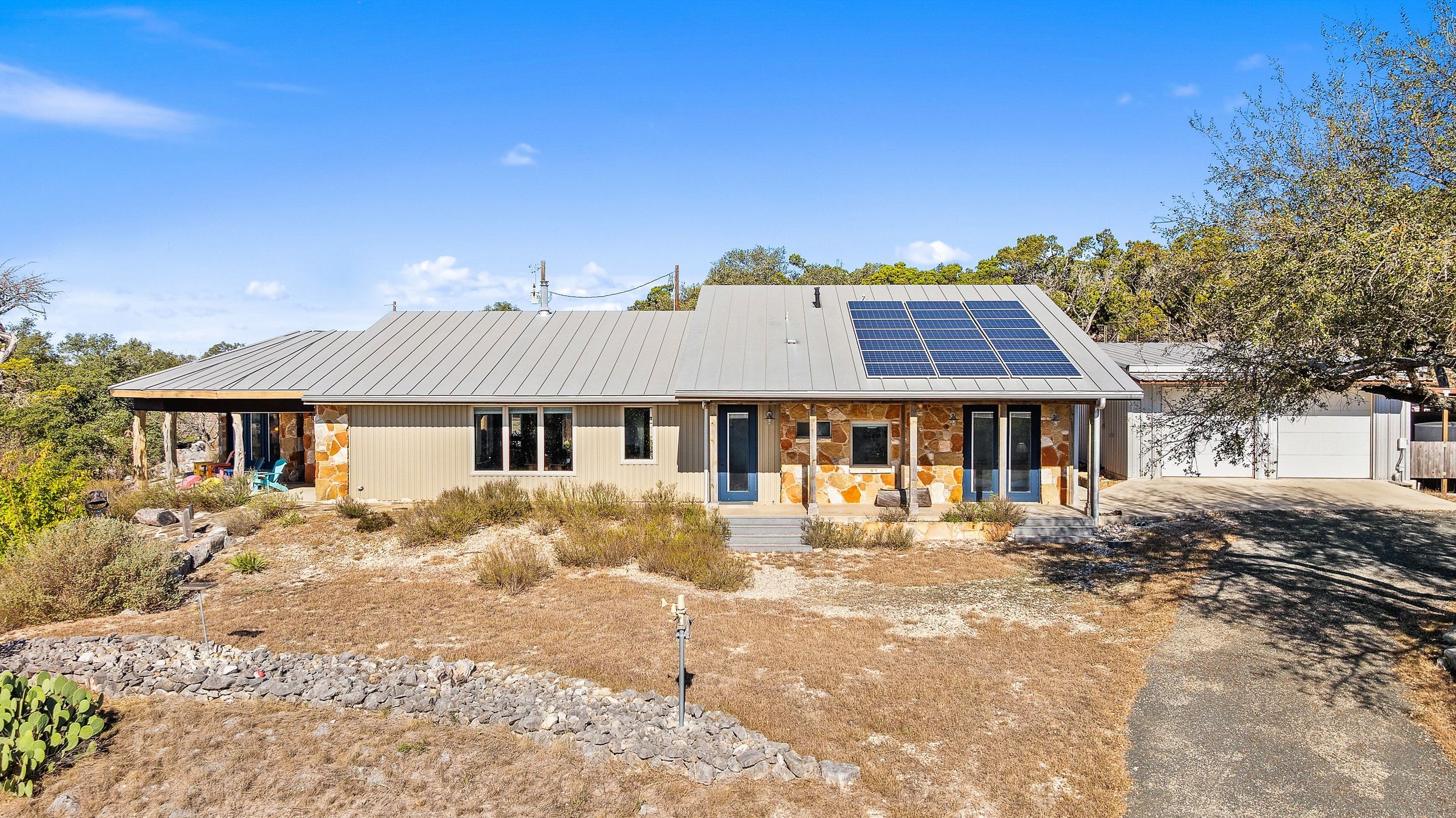 A rustic-modern house with a metal roof and solar panels, surrounded by a dry yard with sparse vegetation and a few trees, under a clear blue sky.