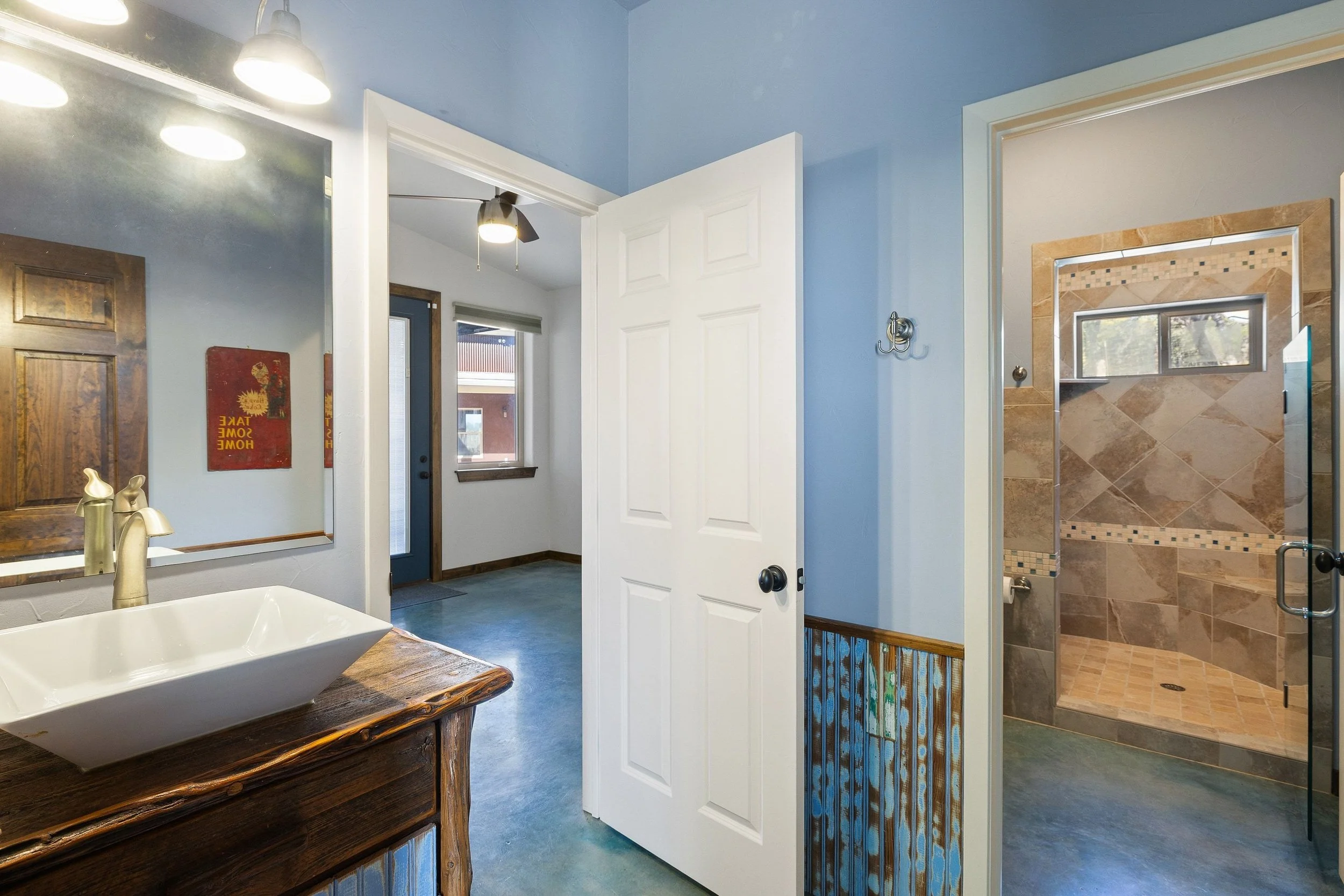 Guest bathroom with a wooden vanity, rectangular white sink, large mirror, and a door leading to a walk-in tiled shower with a window.