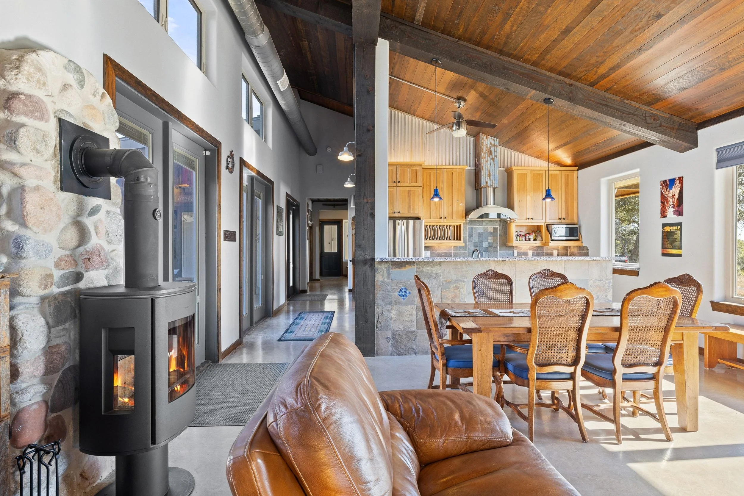 Living room with a wood stove and a leather sofa, dining table with six chairs, open kitchen with wooden cabinets, pendant lights, and large windows with natural light.