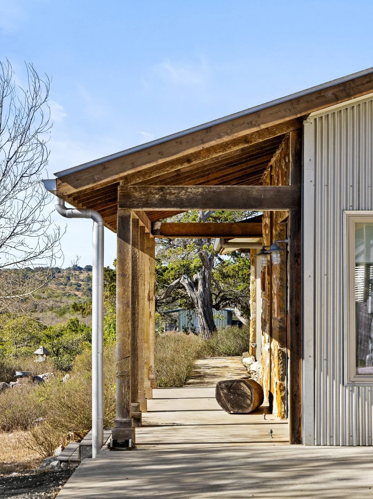 Exterior view of a rustic house with a corrugated metal wall on the right and a wooden porch with logs and beams. The porch is shaded by an overhanging roof, and the landscape includes trees, bushes, and distant hills under a clear sky.