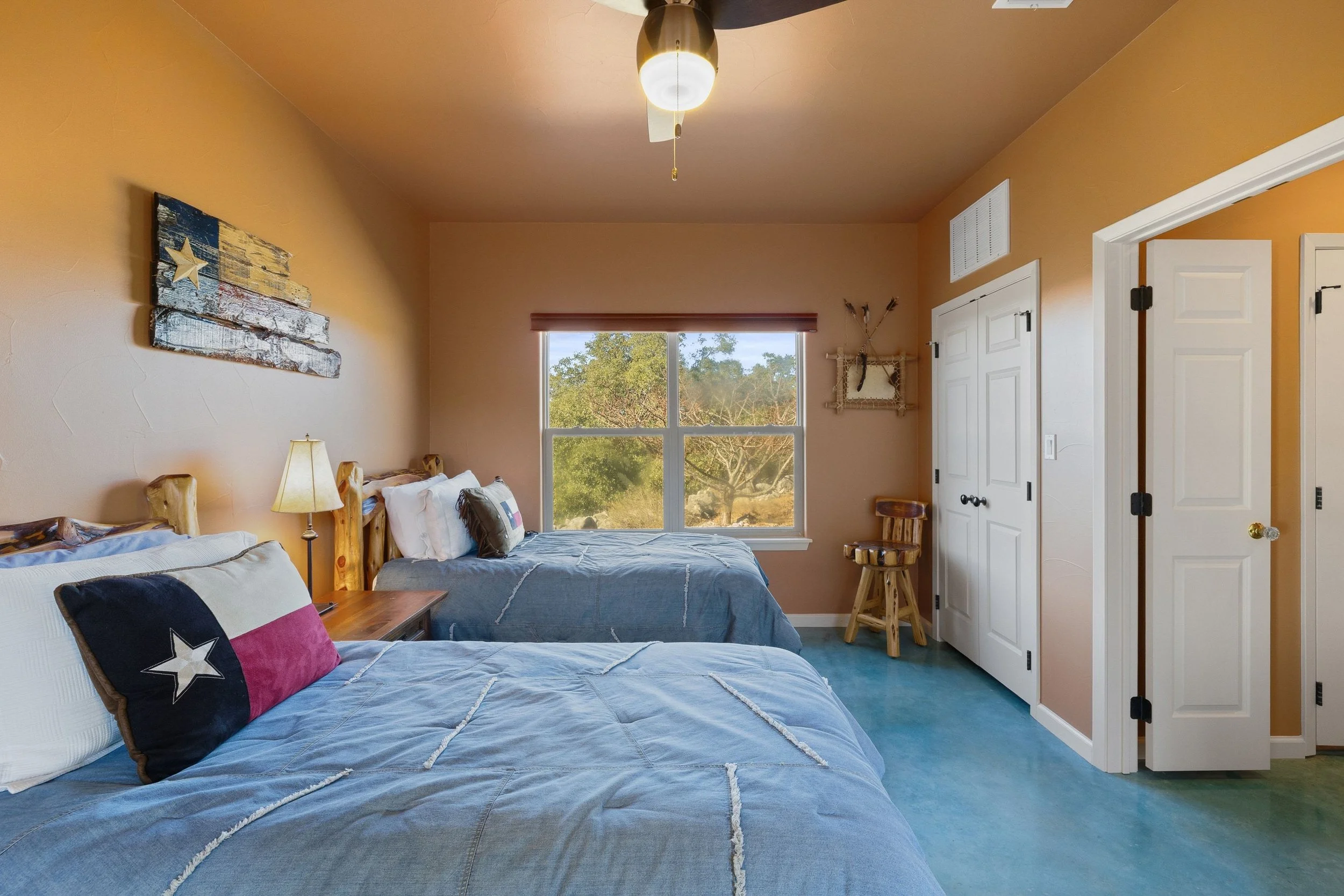 A bedroom with two beds, a window showing outdoor trees, a wooden chair, and rustic decor including a wall hanging made of arrow and a Texas flag-style pillow on one bed.