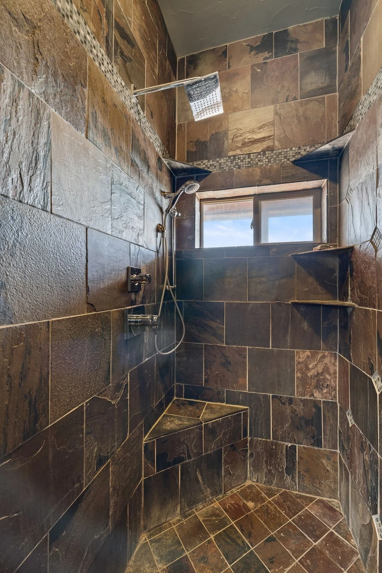A walk-in shower with dark brown stone tiles on the walls and floor, a window near the ceiling, a rain shower head, a handheld shower head, and built-in wall shelves.
