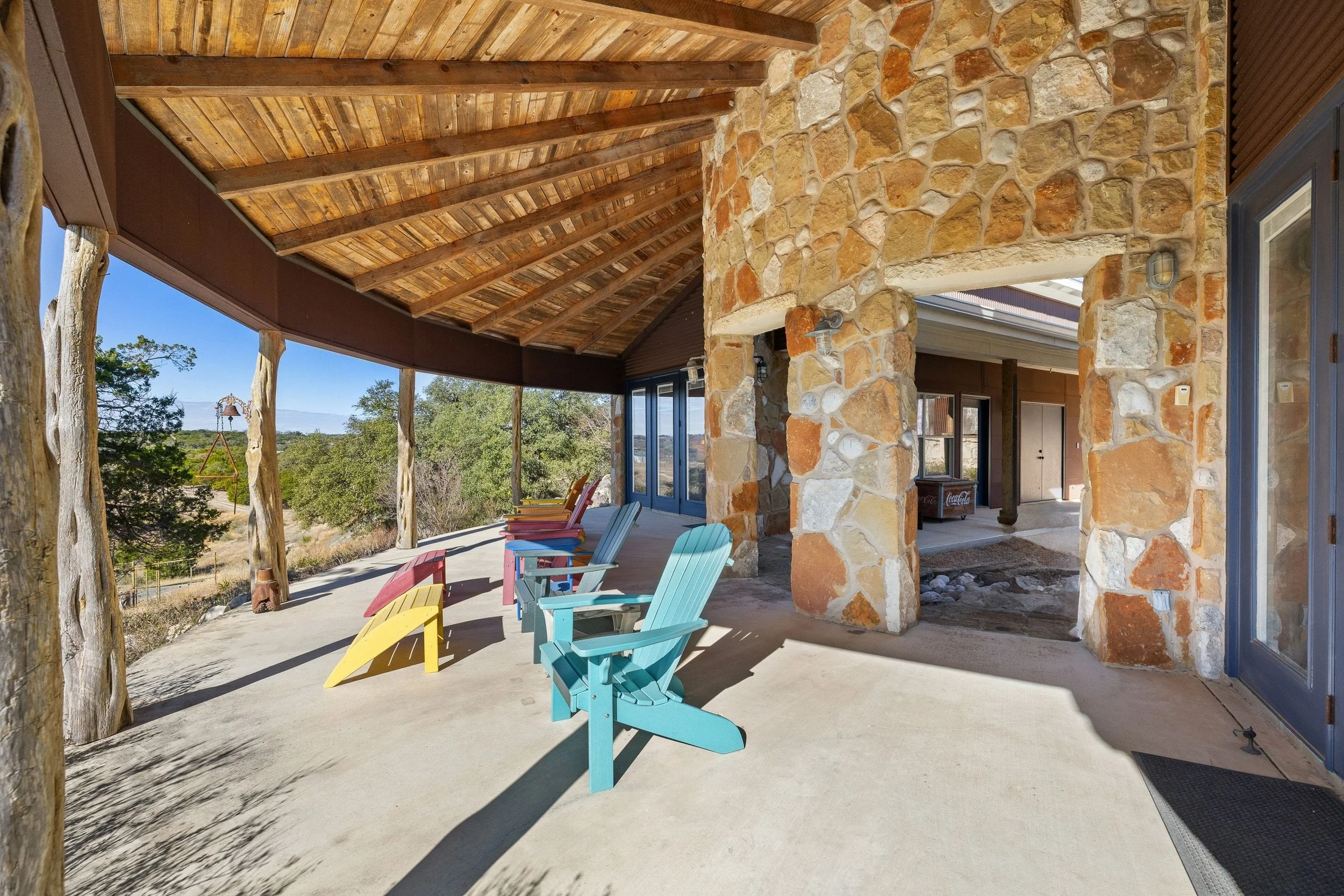 Colorful Adirondack chairs on a spacious covered porch with stone columns, overlooking a natural landscape with trees and a clear blue sky.