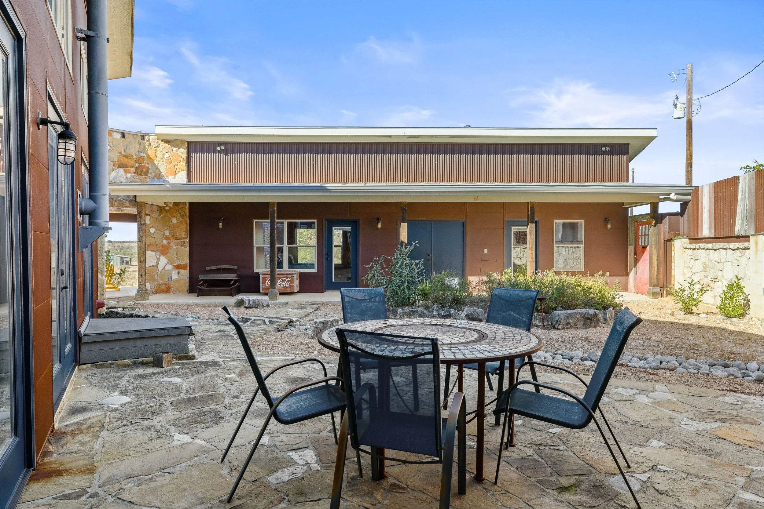 Outdoor courtyard area with a round table and four chairs, stone pathway, and a modern house in the background with stone and wood exterior walls, windows, and a sliding glass door.