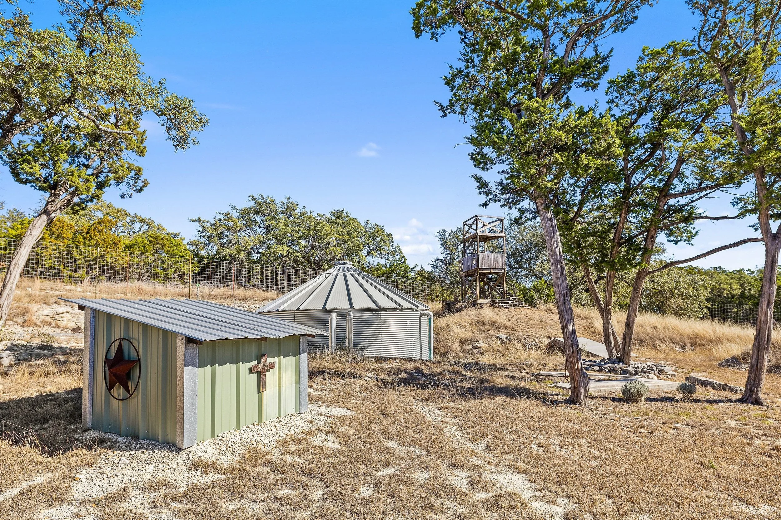 Outdoor scene with a small shed, a round metal water tank, and a wooden observation tower among trees under a clear blue sky.