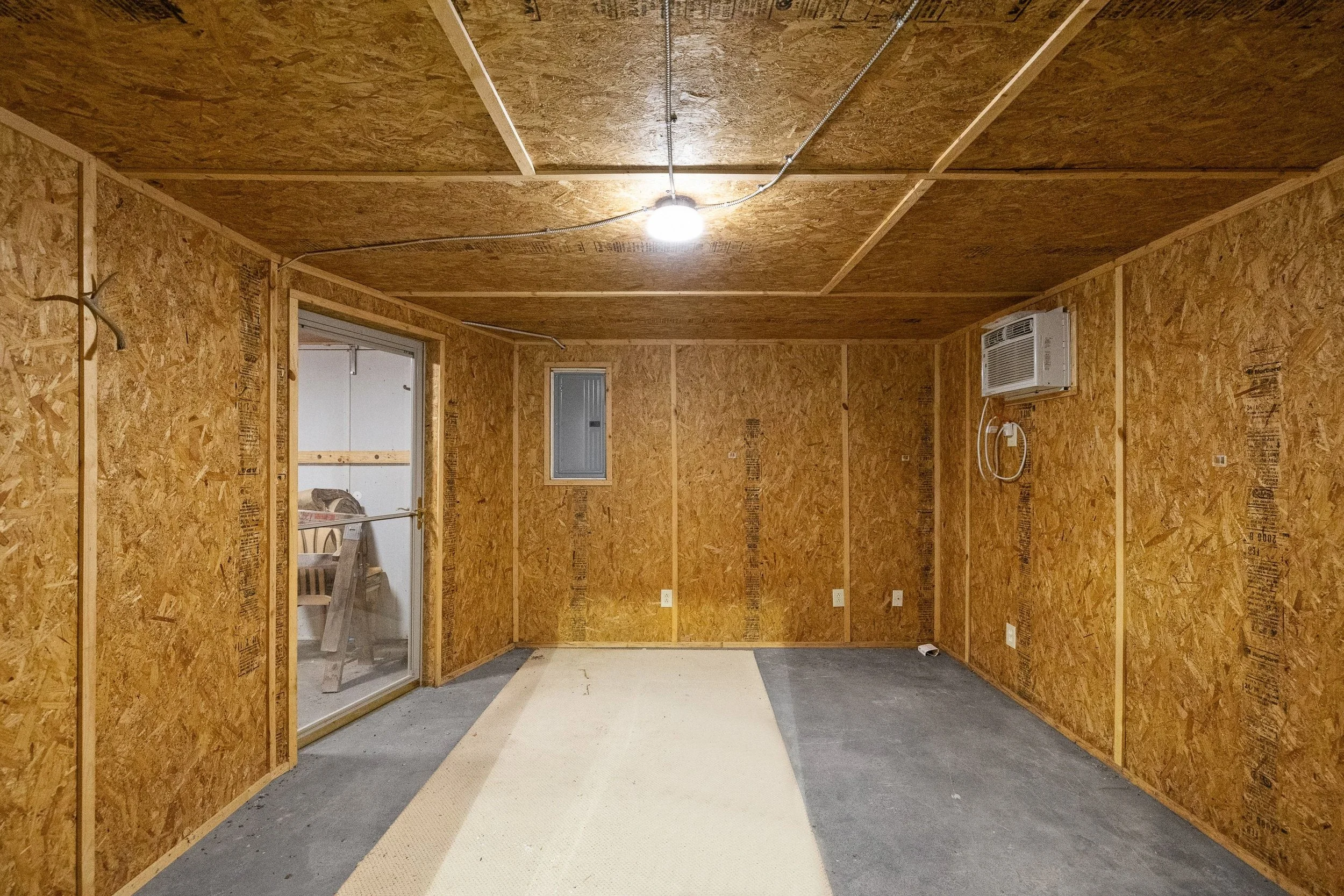 Empty room with unfinished wooden walls and ceiling, gray concrete floor, small window, air conditioning unit, and sliding glass door leading to an outdoor space.