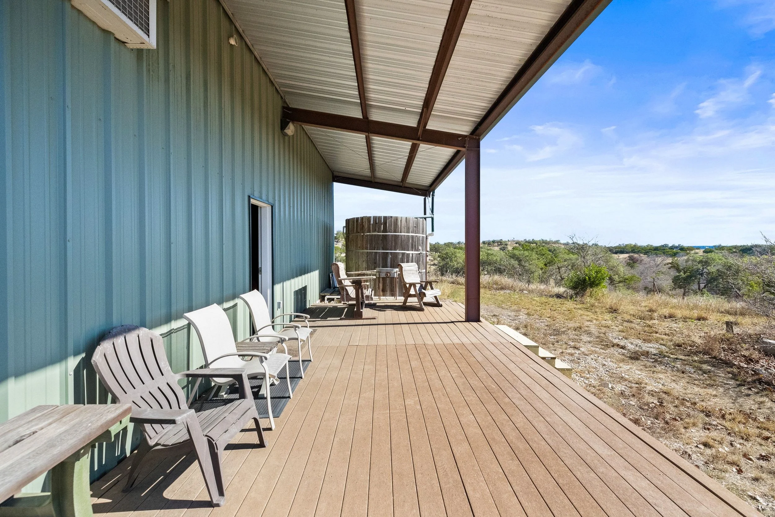 Outdoor porch of metal barn with several white chairs along a green building, overlooking a landscape with bushes and a blue sky.