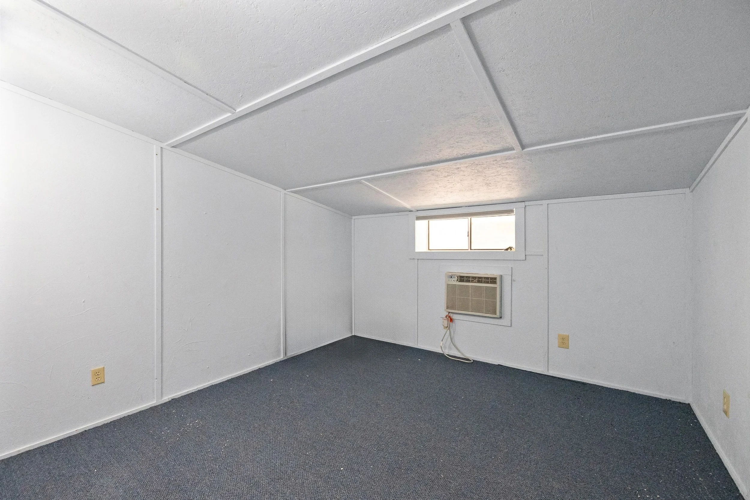 Empty small room with white walls, a gray carpeted floor, a small rectangular window, and a wall-mounted air conditioning unit.