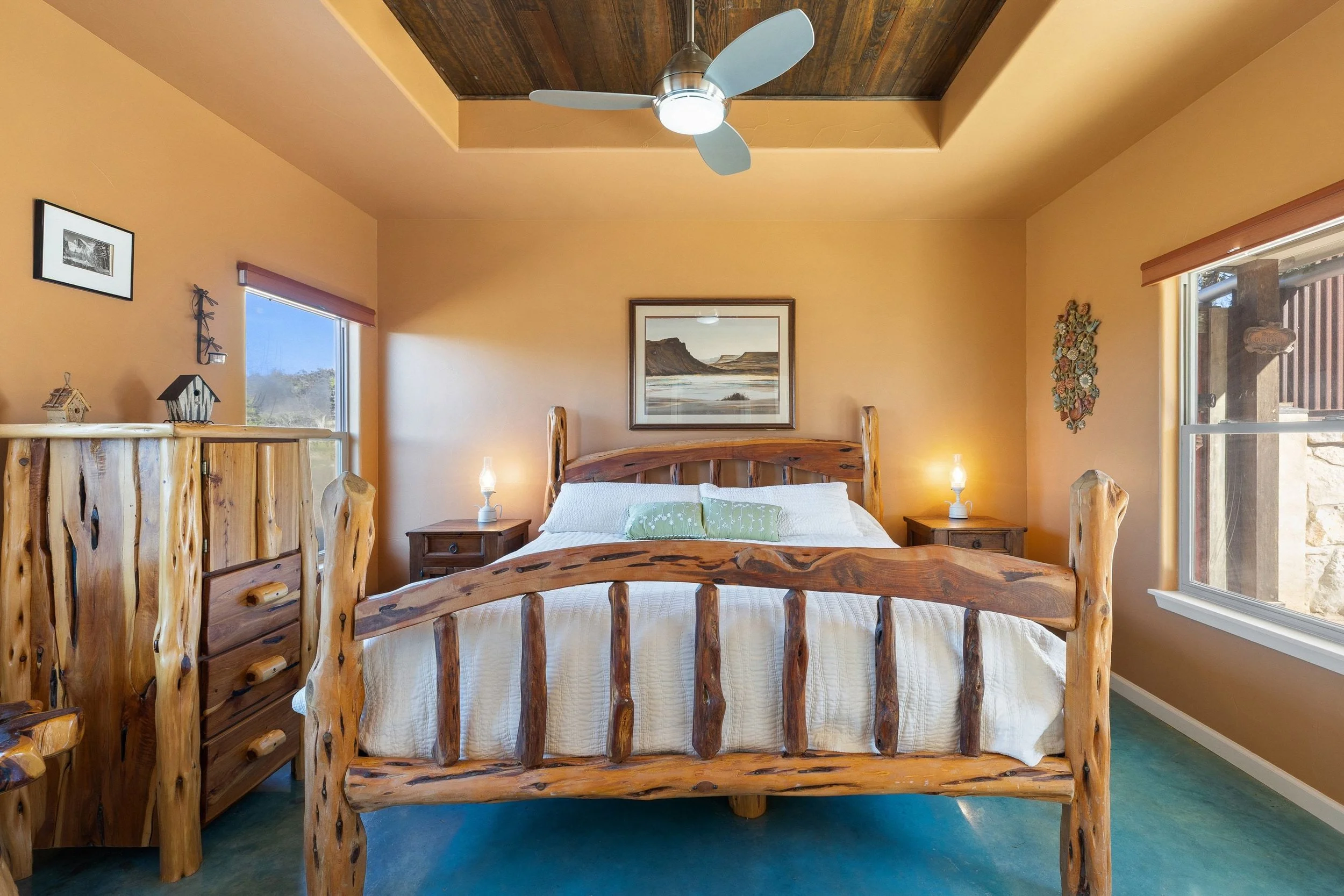 Cozy bedroom with rustic wood furniture, including a large bed with a wooden headboard and footboard, two bedside tables with lamps, a dresser, and framed artwork on the walls, illuminated by natural light from windows and a ceiling fan.