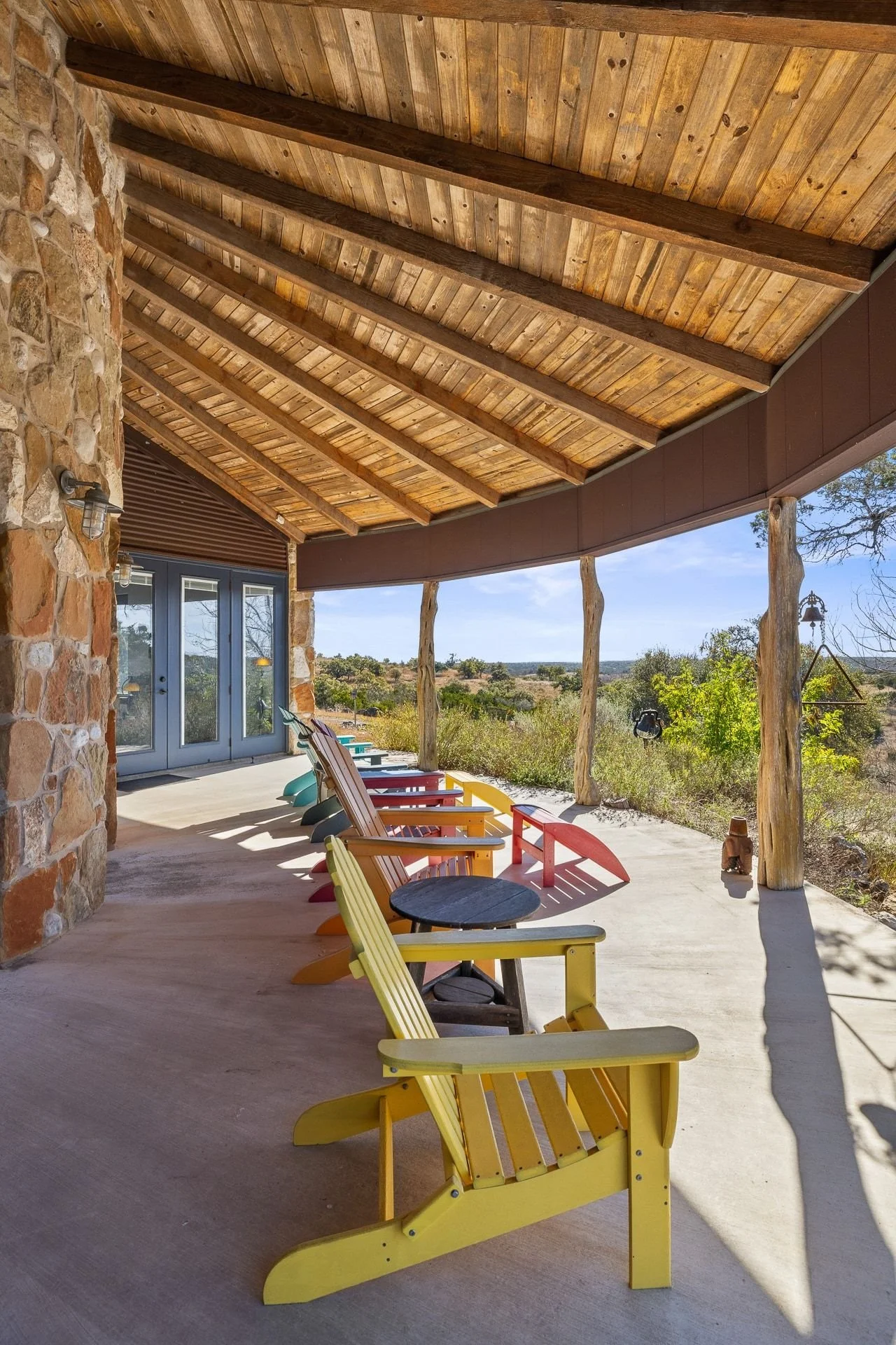 A covered porch with colorful Adirondack chairs and small tables, overlooking a scenic Texas Hill Country landscape with trees and a blue sky.