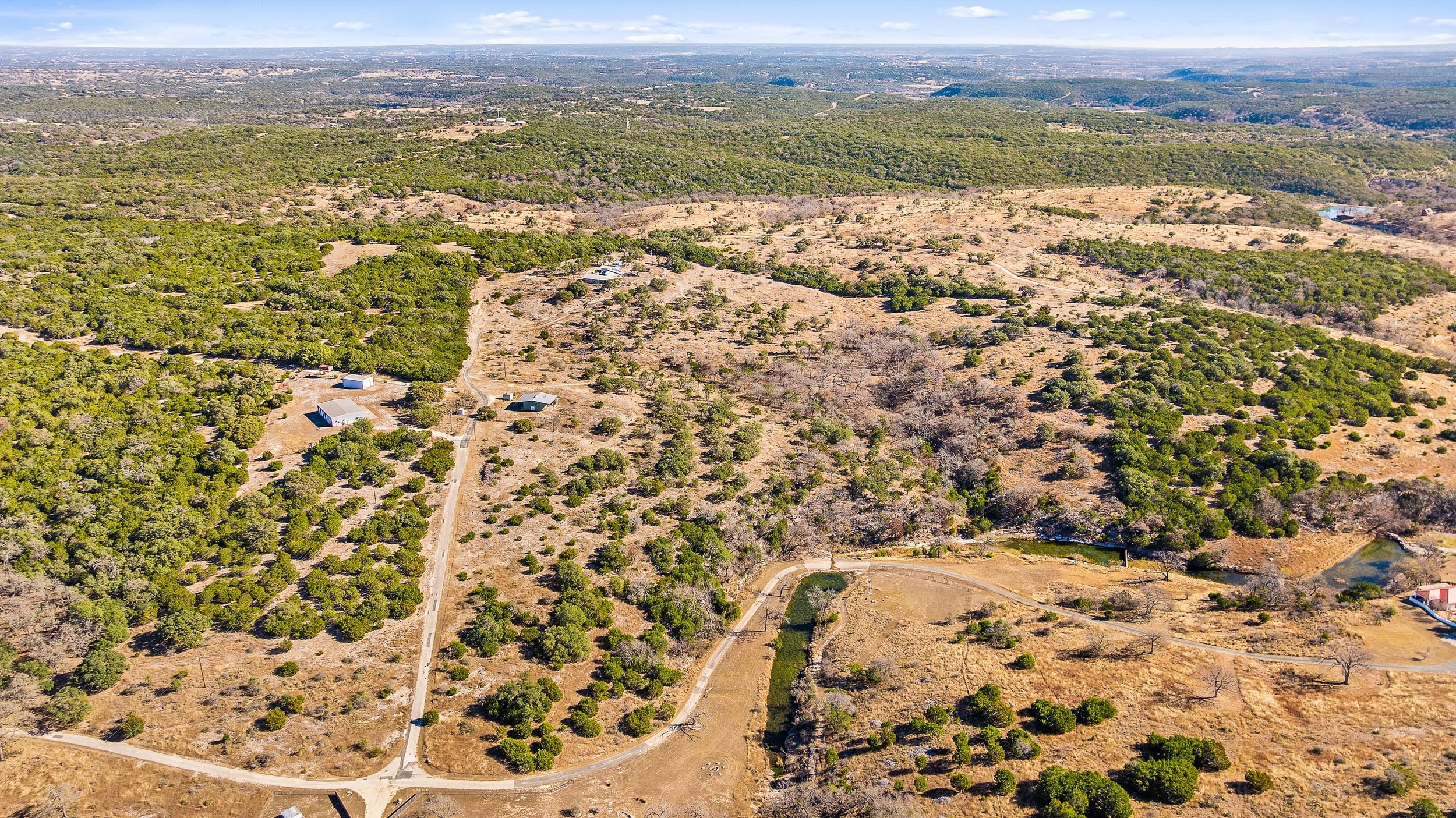 Aerial view of a semi-arid landscape with scattered trees, dirt roads, and few small buildings, stretching to a distant horizon with a mix of forested and open areas.