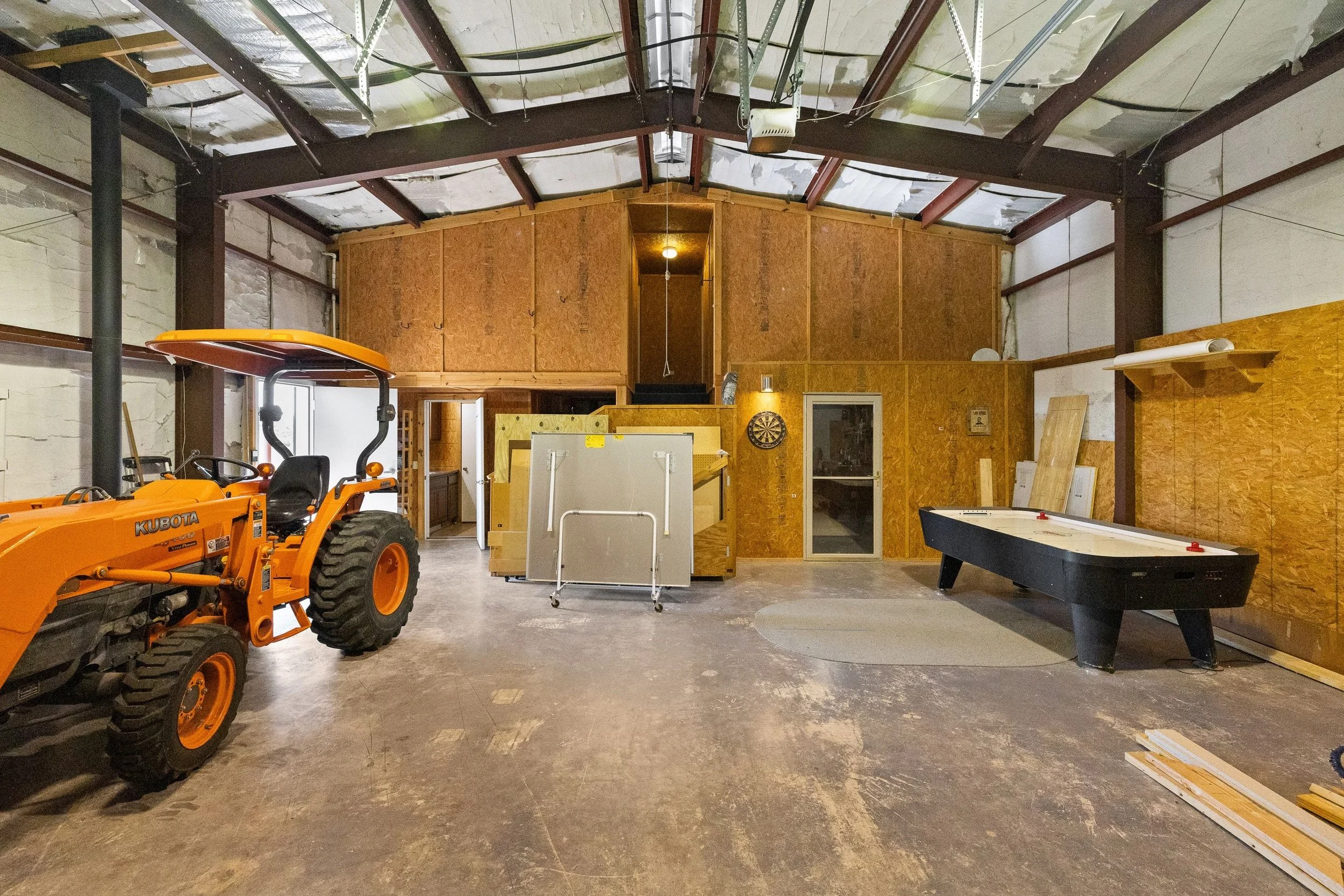 An unfinished indoor garage or workshop with a small orange Kubota tractor on the left, a dartboard on the back wall, an air hockey table on the right, and construction materials and furniture scattered around.