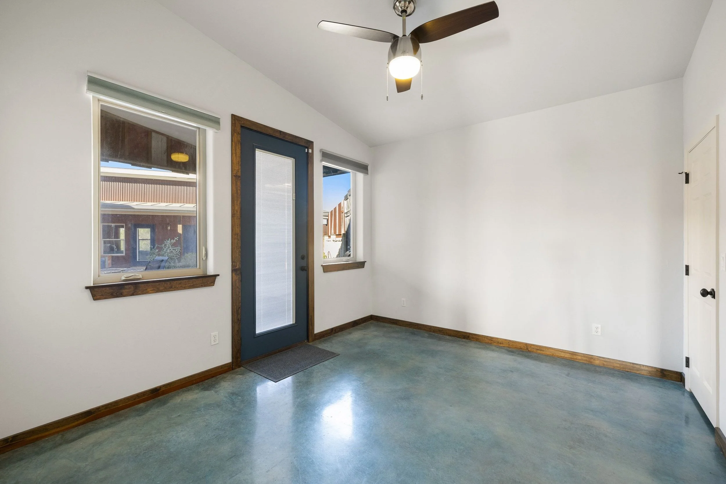 Guest room with white walls, two windows, a dark blue door with a glass panel, and a ceiling fan with wooden blades.