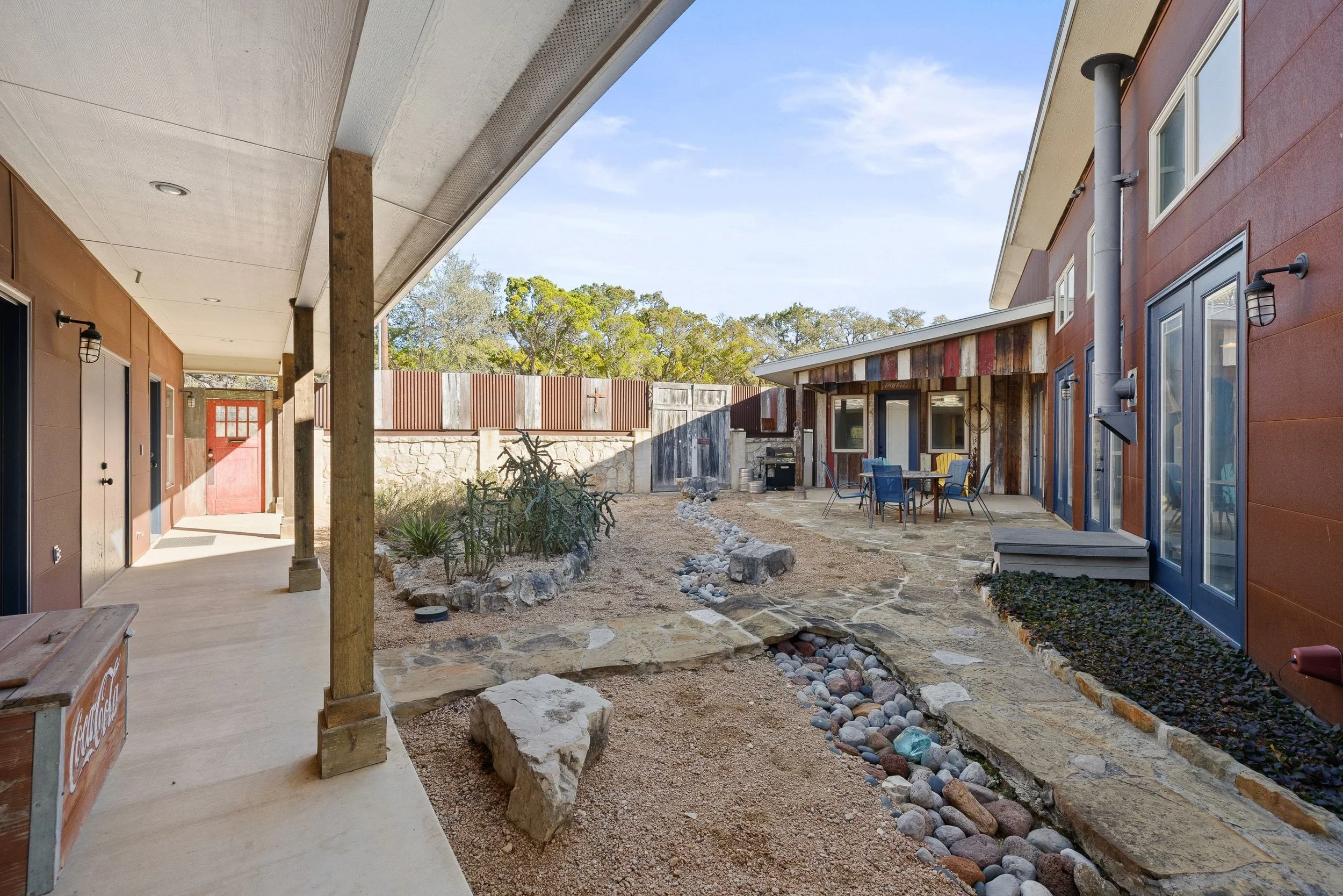 An outdoor patio area with a pathway made of stone, surrounding plants and rocks, adjacent to a house with a covered porch on the left and a patio with outdoor furniture on the right. The house has a mixed material exterior with wood siding and large