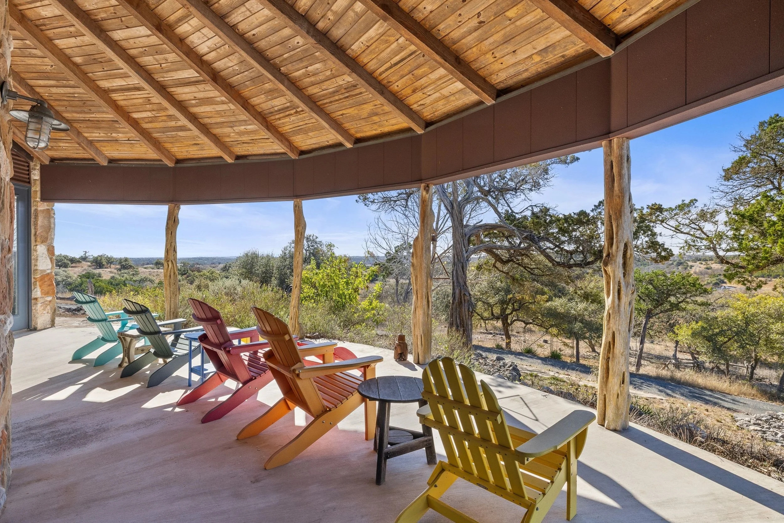 Outdoor patio with colorful Adirondack chairs and small table, overlooking a scenic Texas Hill Country landscape with trees and open land.