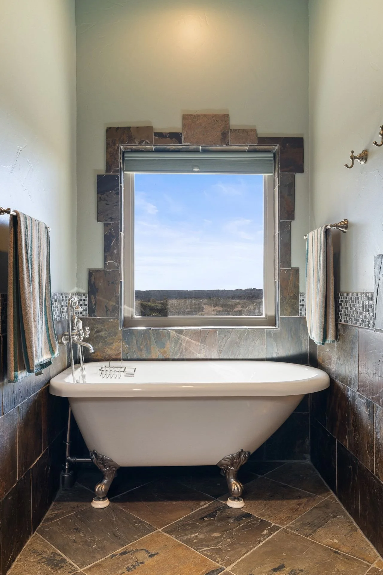 A vintage clawfoot bathtub with ornate metal feet is placed in front of a window overlooking a landscape. The bathroom has dark marble tiles on the floor and similar tiles on the lower walls, with a light-colored painted wall above. There are towels 