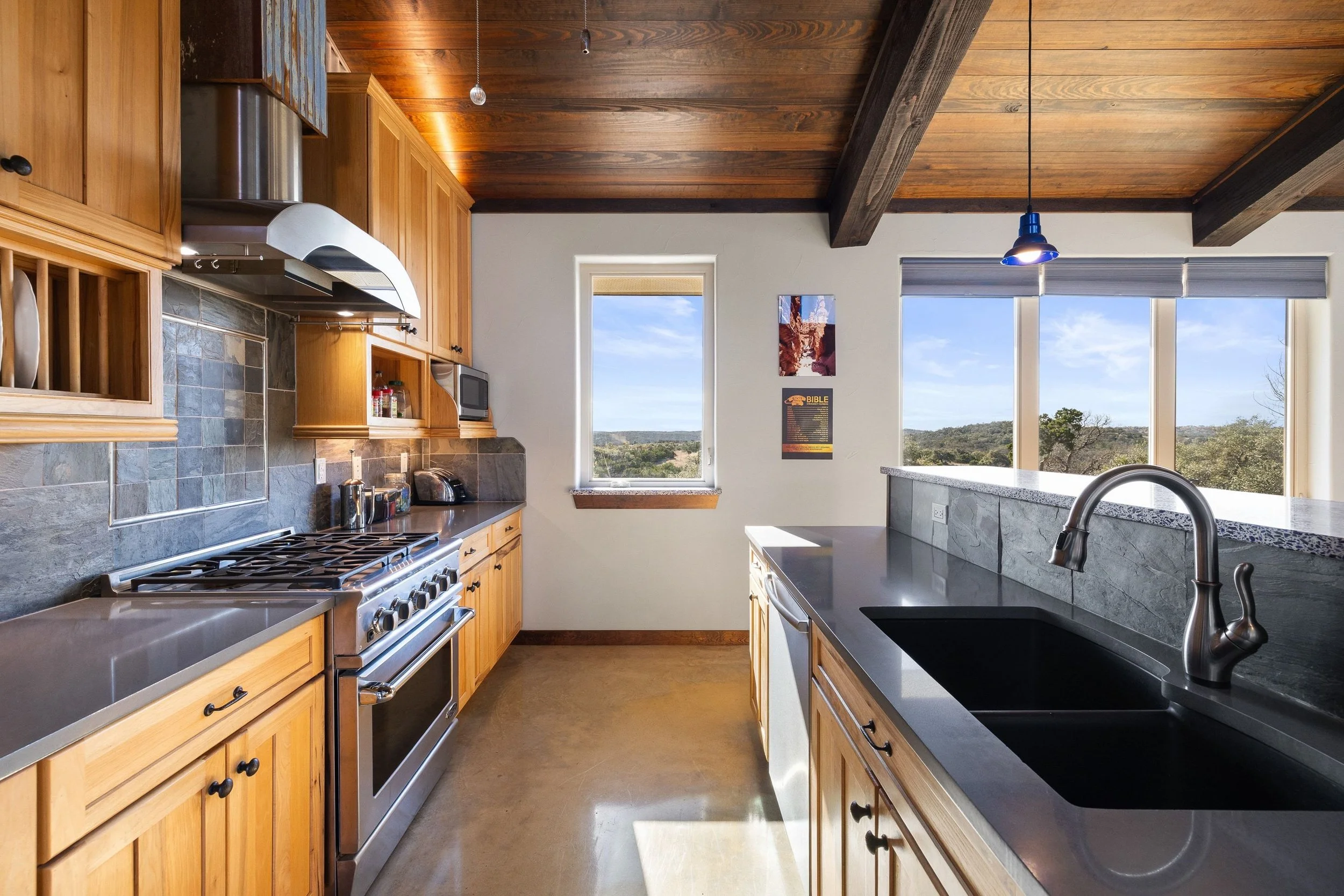Kitchen with wooden cabinets, stainless steel stove, black countertops, double sink, large windows showing an outdoor landscape, wooden ceiling with beams, and a pendant light.