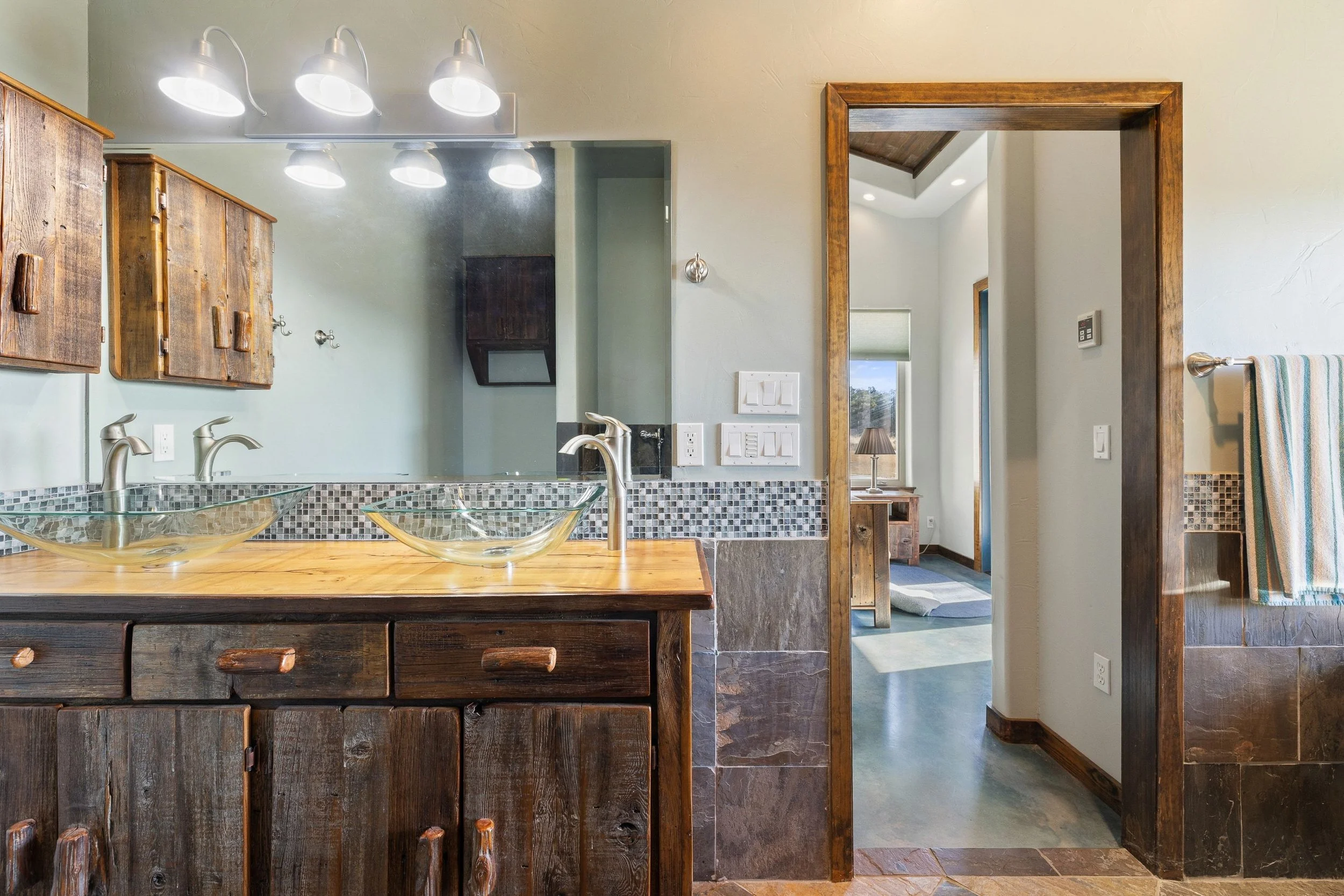 Bathroom with a double vanity featuring glass vessel sinks, wood cabinets, a large mirror, and modern light fixtures. There is a doorway leading to a room with a window and a side table with a lamp.