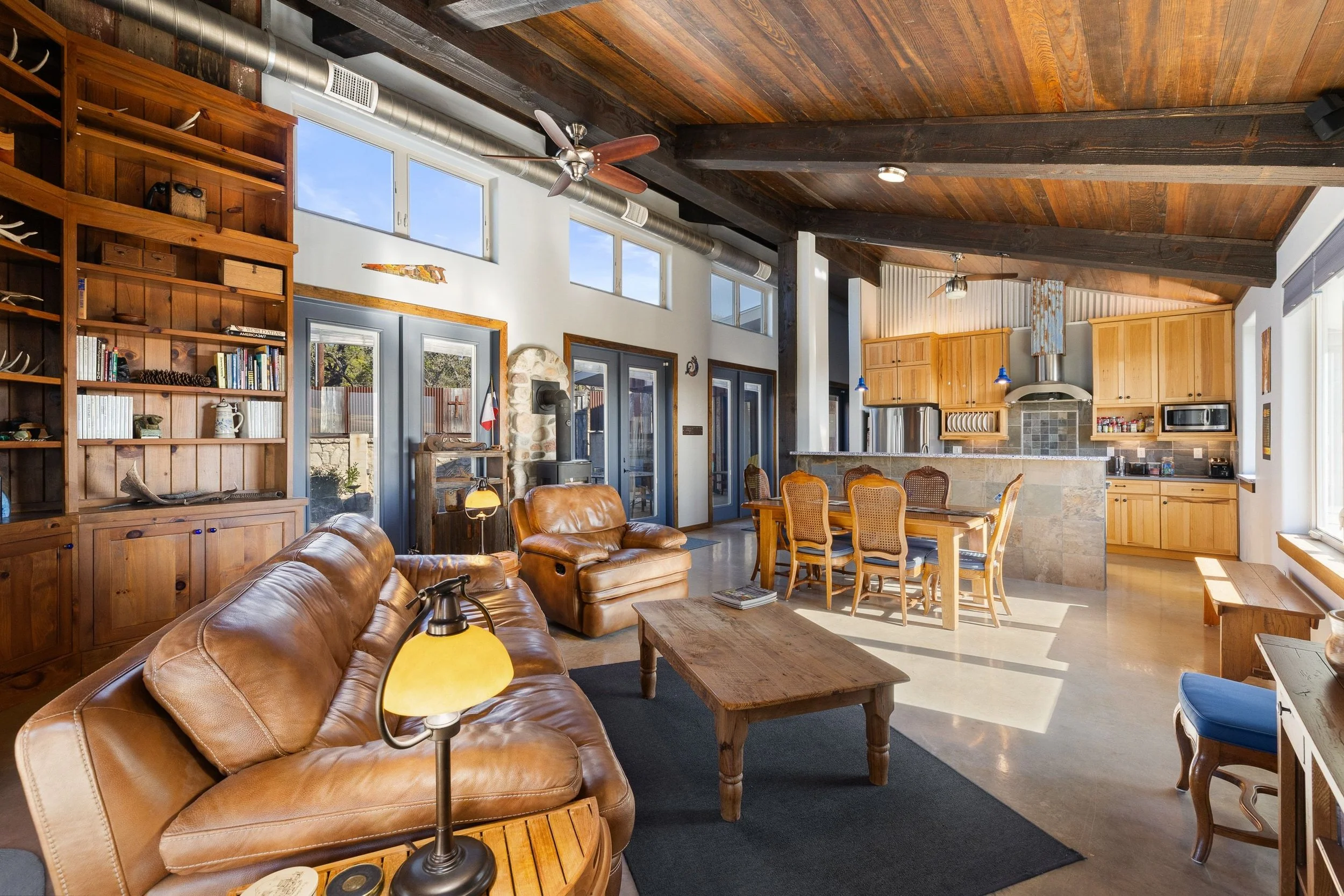 Open-concept living space with leather sofas, a wooden coffee table, a dining table with chairs, and a kitchen with wooden cabinets, a stone island, and stainless steel appliances. Large windows let in natural light.