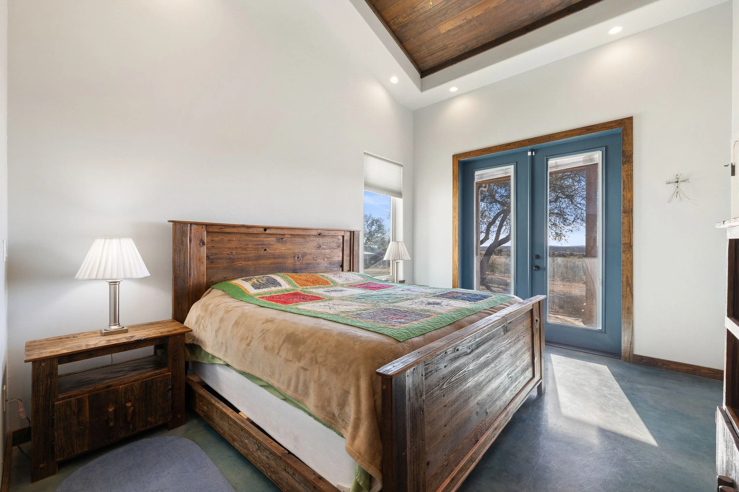 A bedroom with a wooden bed frame, colorful quilt, and two bedside lamps. It has white walls, a window, and French doors leading outside with a view of trees and a landscape.