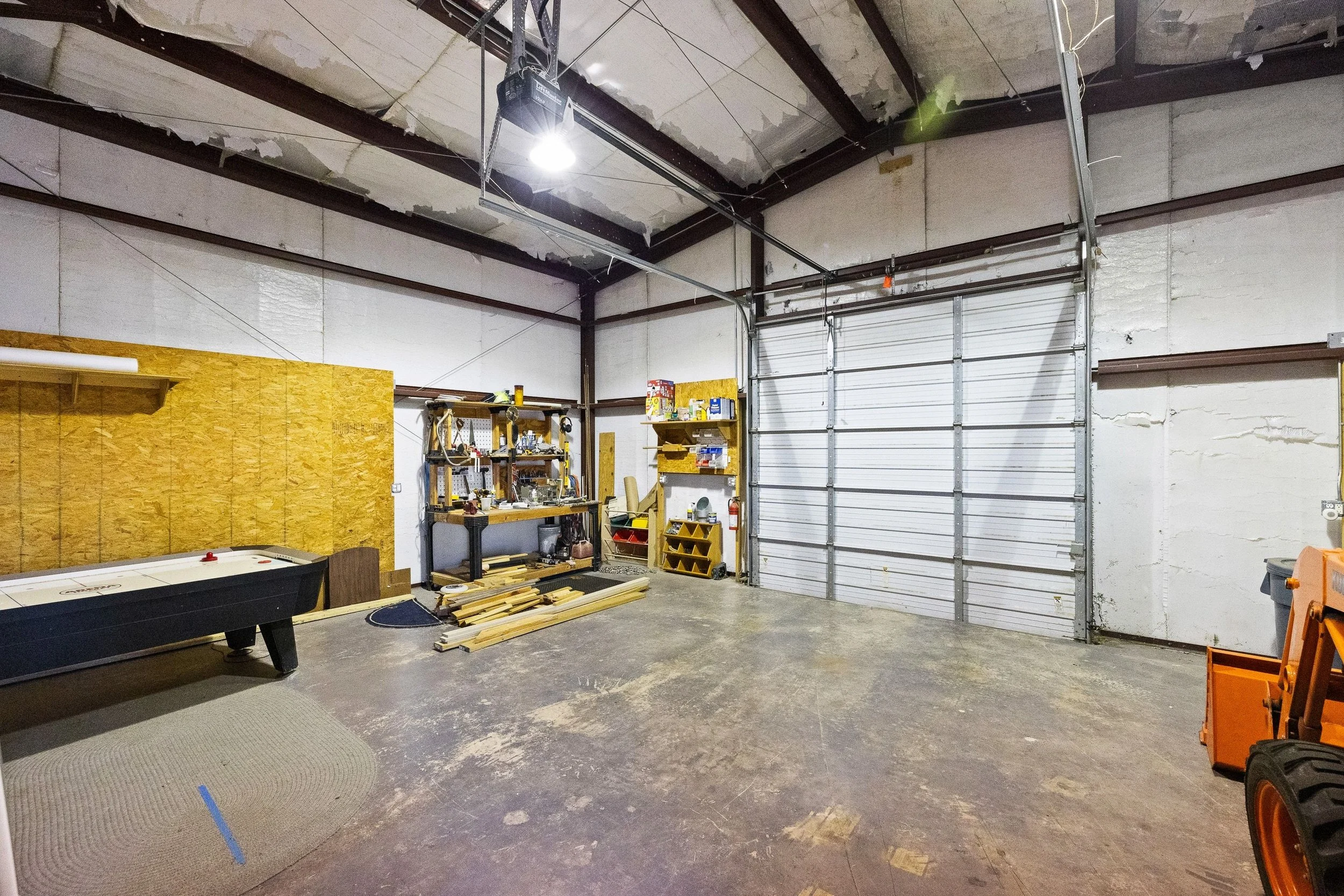 Empty garage or workshop space with a garage door and shelving with tools and supplies; a shuffleboard table and some wooden planks on the floor.