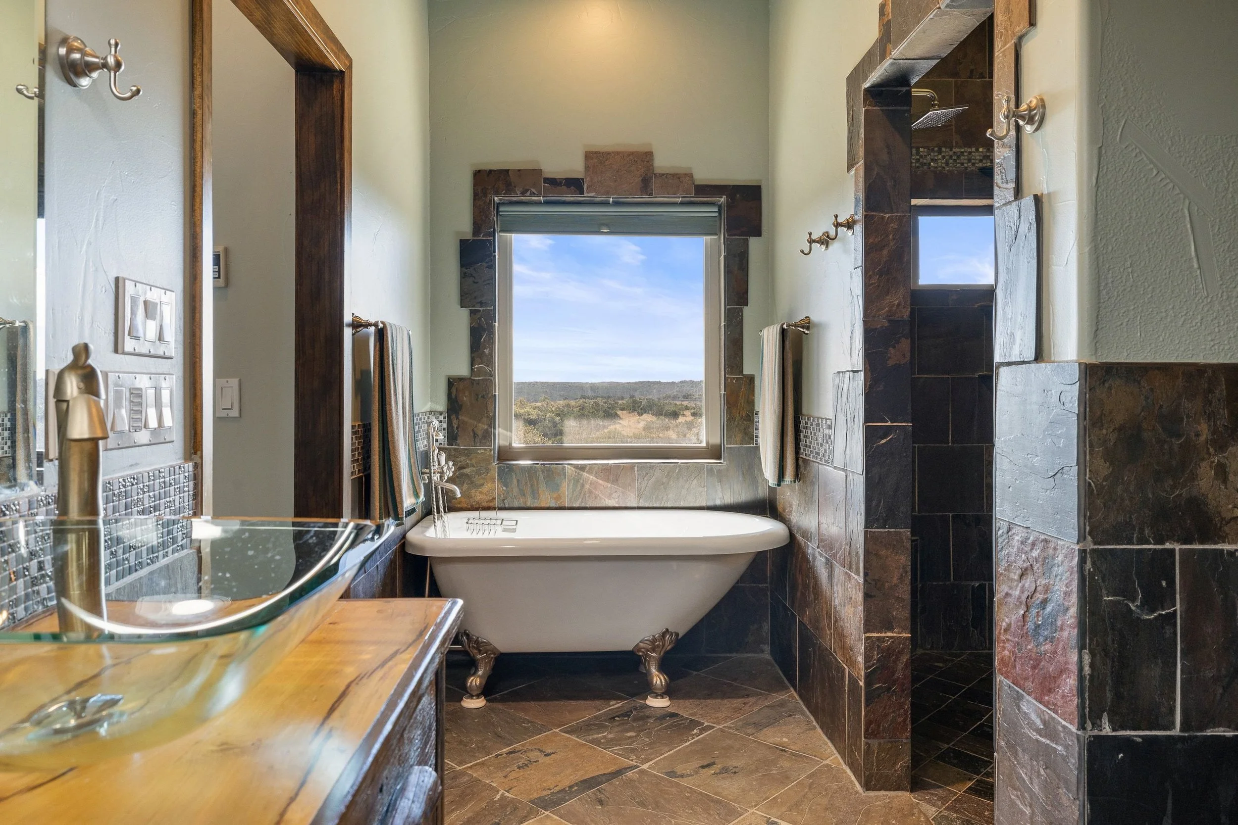 Bathroom with a vintage clawfoot bathtub under a large window, tiled walls with mosaic accents, stone floor, and a glass vessel sink on a wooden vanity.