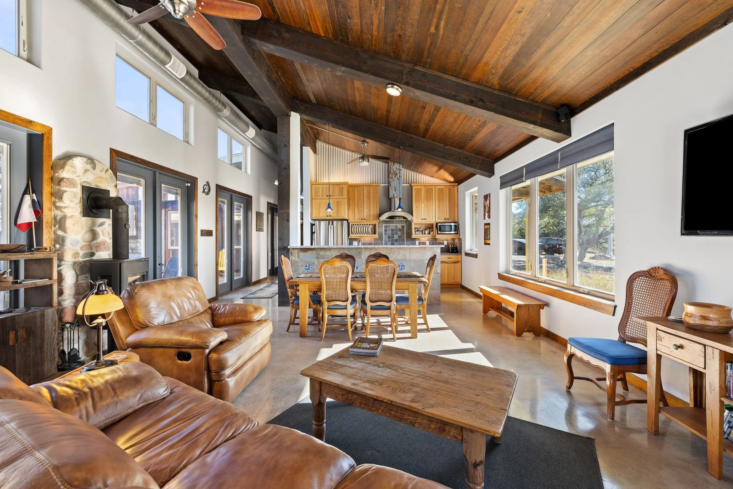 Open-concept living room and kitchen with wooden ceiling, leather sofas, dining table with chairs, large windows, and a wood stove.