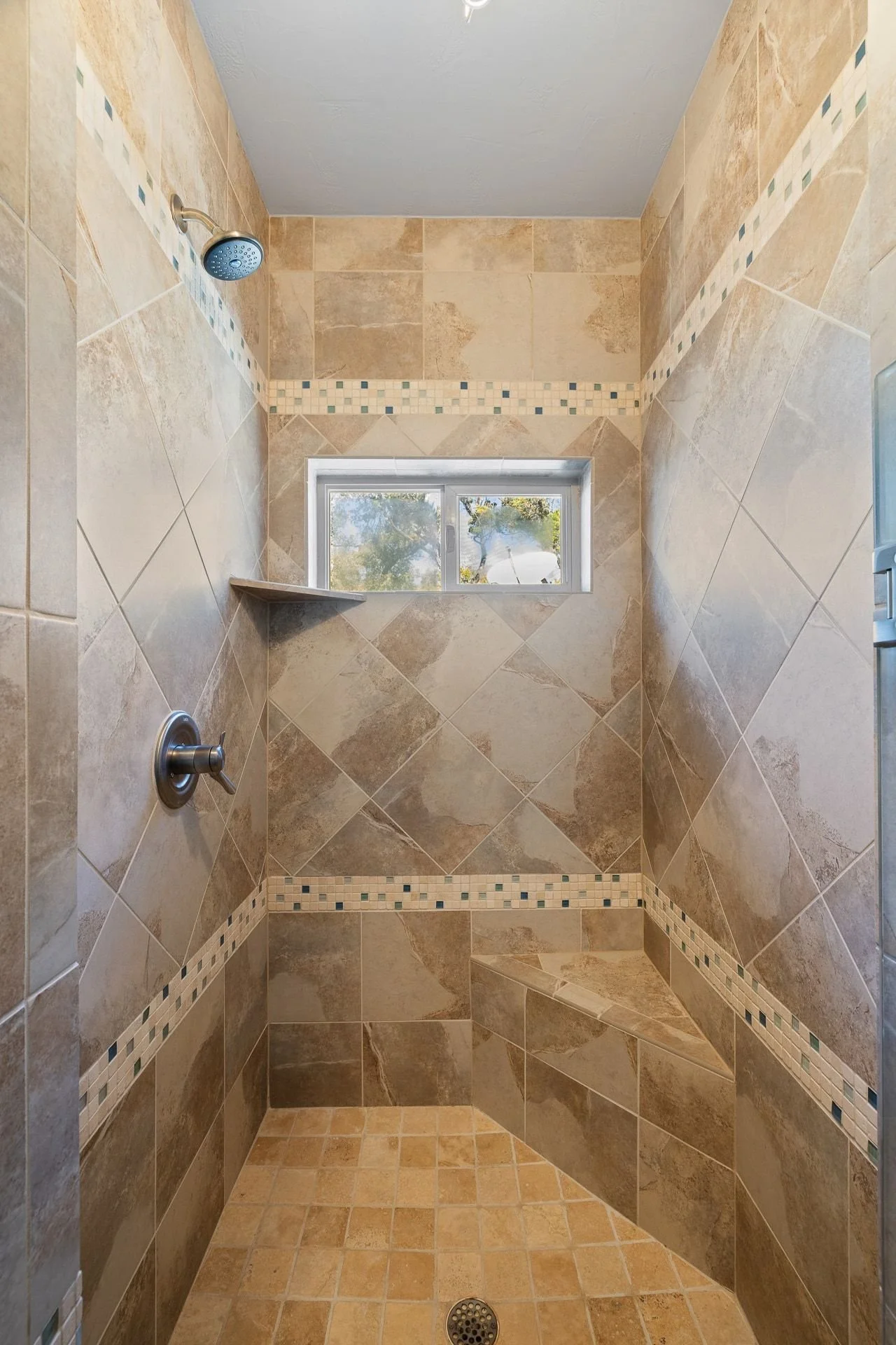 A walk-in shower with beige and brown tiled walls and floor, a small horizontal frosted window, a corner shelf, a showerhead on the left wall, and a drain on the floor.
