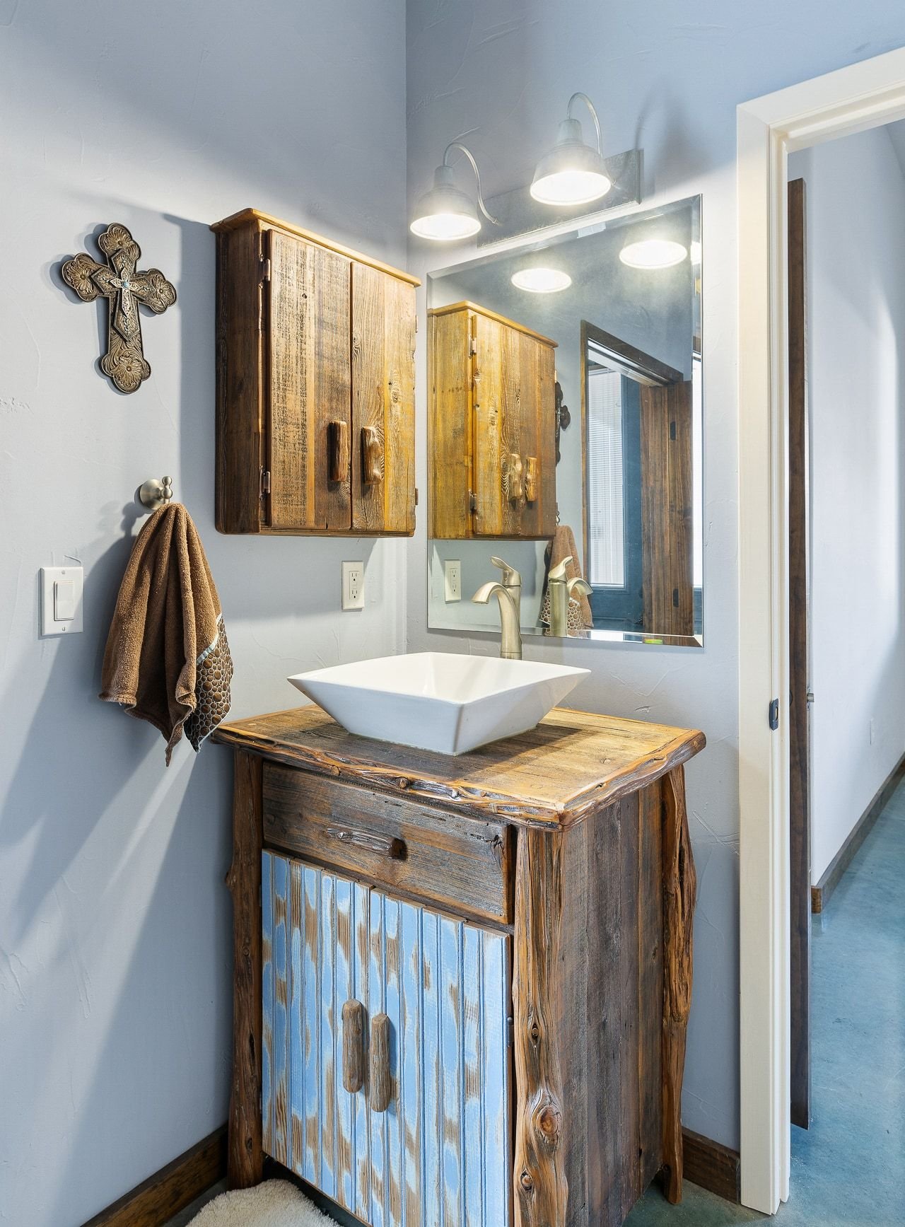 Rustic bathroom vanity with a square vessel sink, wooden cabinets, and a large mirror with three light fixtures. A brown towel hangs on a metal hook, and a decorative cross is on the wall.
