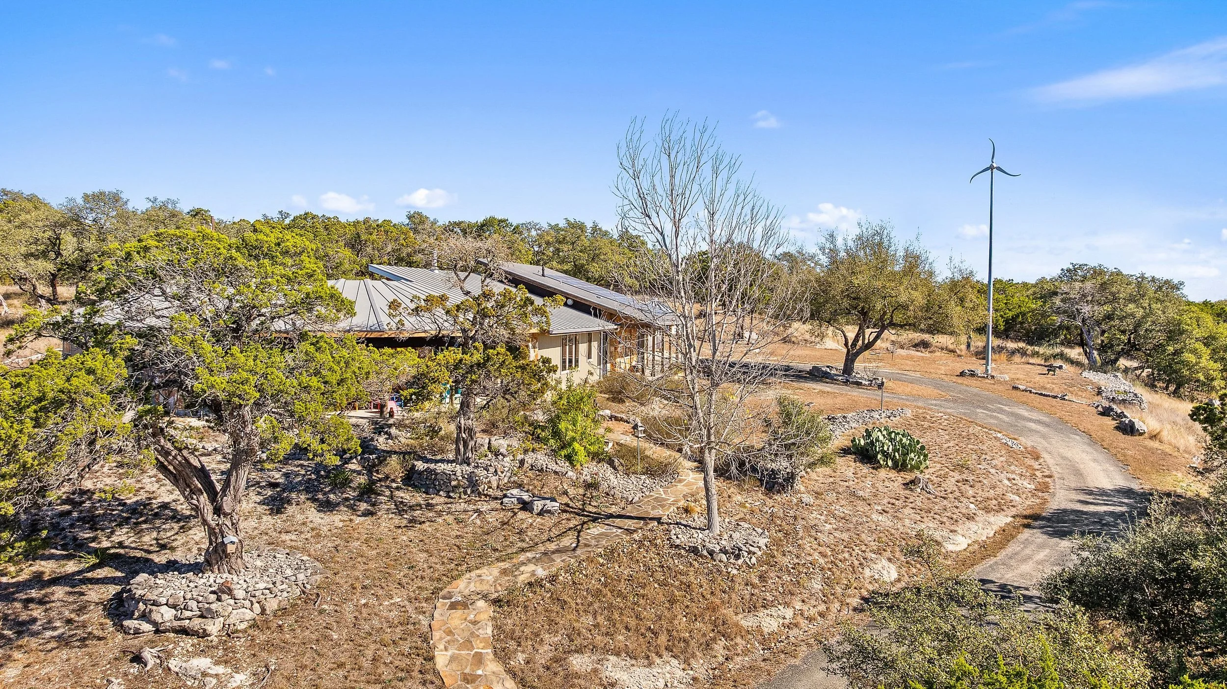 A house on a hill surrounded by trees with a wind turbine in the background, a dirt road curves around the landscape under a blue sky.