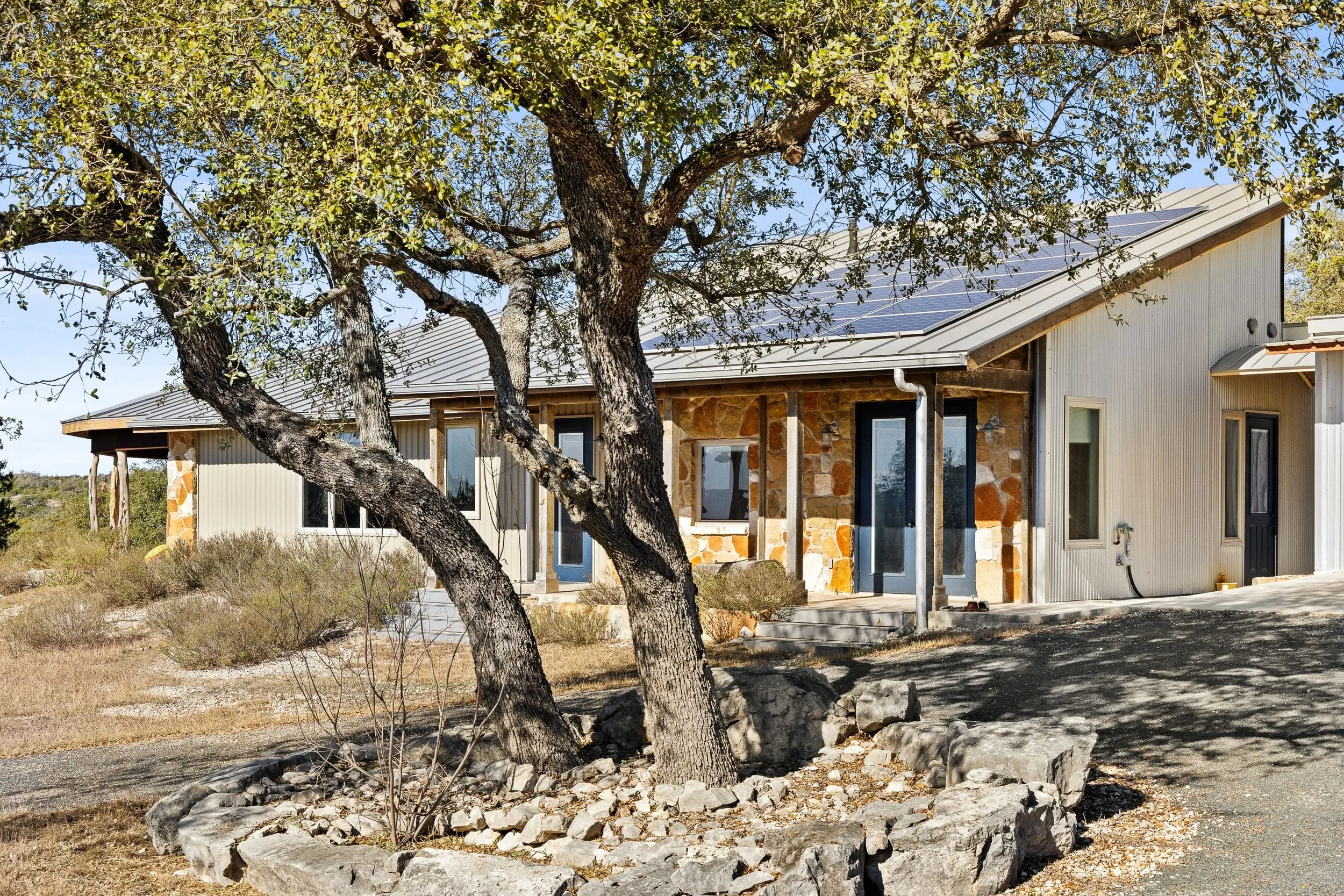A modern, rustic Hill Country home with a metal roof and stone accents, set amid dry landscape with a large tree in the front yard.