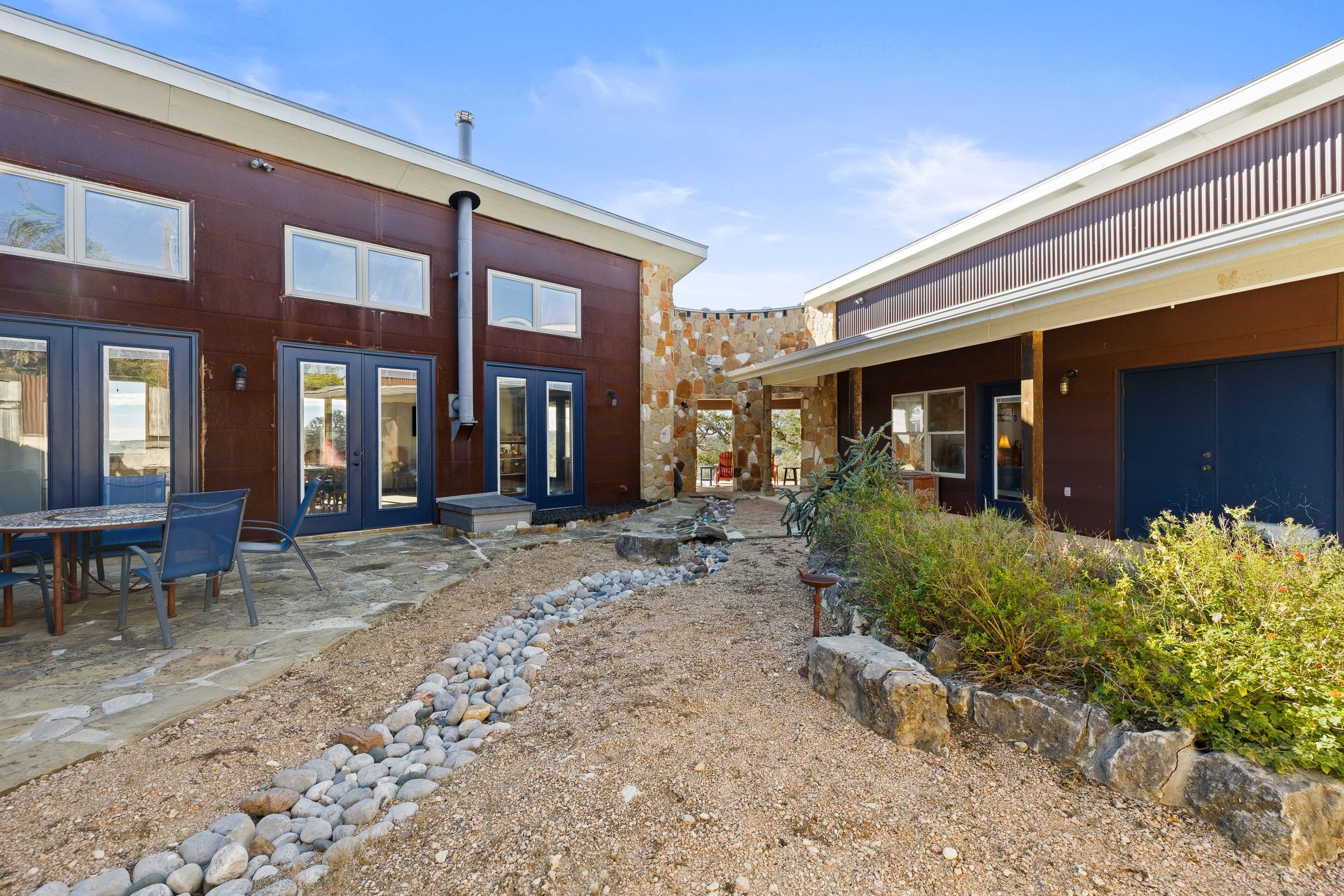 Rustic-modern courtyard with stone pathway, outdoor dining table, and sitting area between two contemporary buildings with brown and beige exteriors under a blue sky.