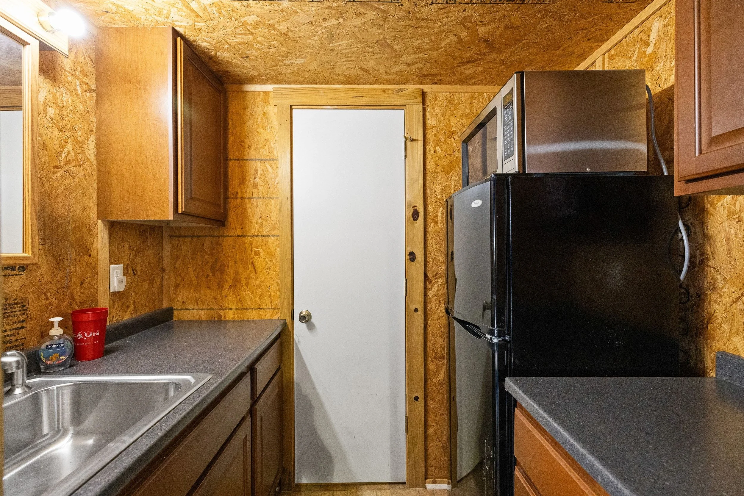 Small kitchen with wood panel walls, a white door, gray countertops, a stainless steel sink, a black refrigerator, a microwave oven, and wooden cabinets.
