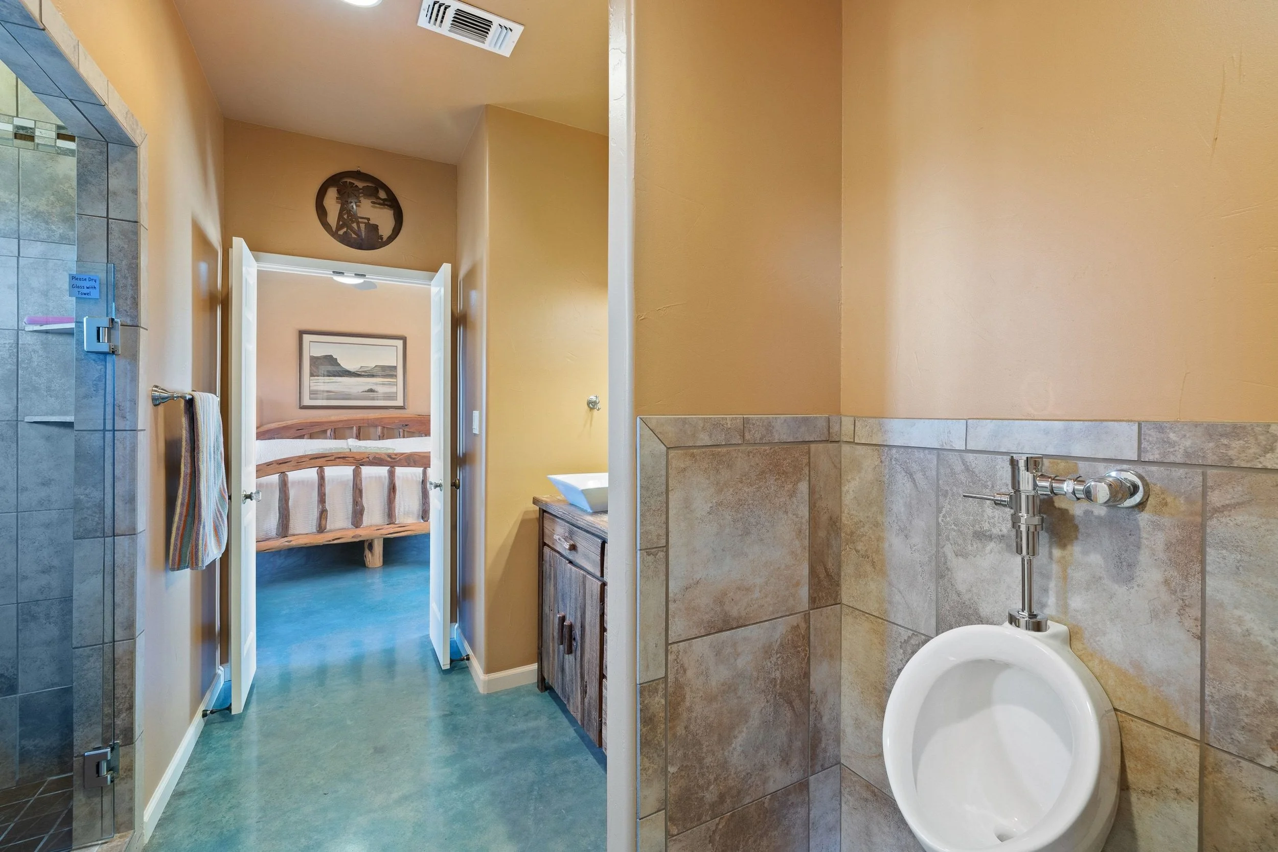 Bathroom with beige walls, a urinal on a tiled wall, a wooden vanity with a sink, a towel hanging, and a doorway leading to a bedroom with a wooden bed frame and framed landscape picture.