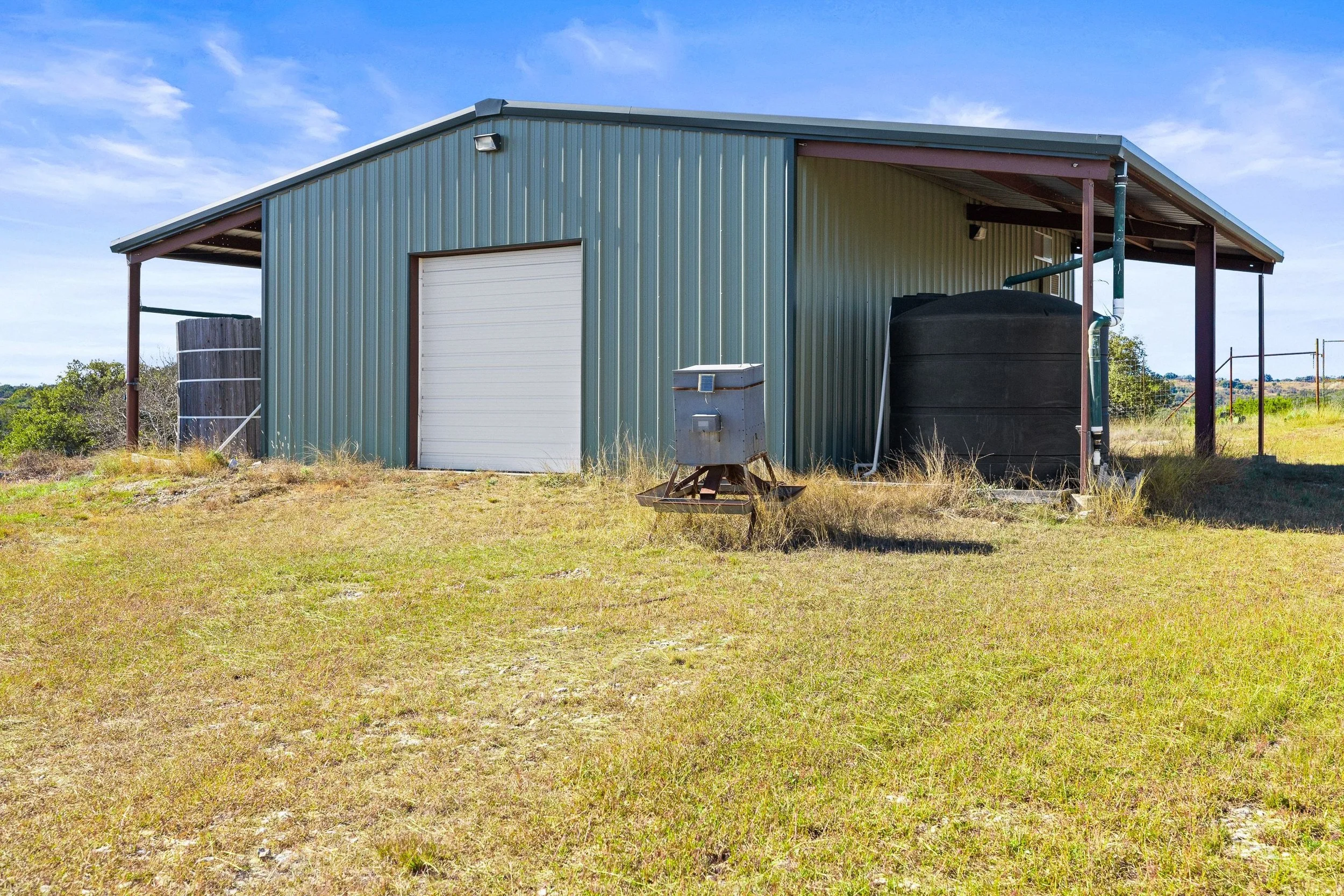 A meta barn with a white roll-up door, situated in a grassy field under a blue sky with scattered clouds. 
