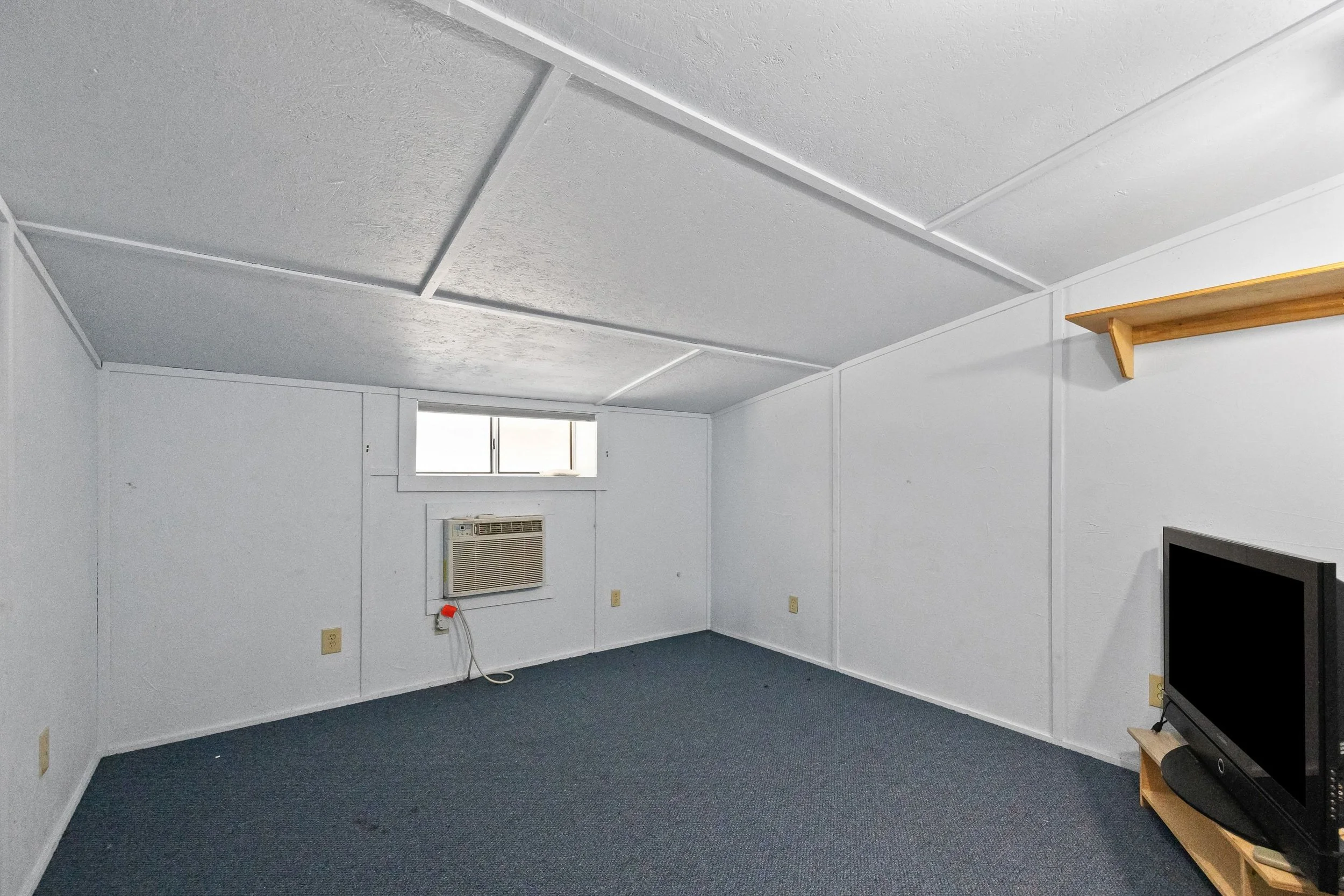 Empty room with white walls and ceiling, a small window near the ceiling, an air conditioning unit, a TV on a small stand, and a wooden shelf on the wall.