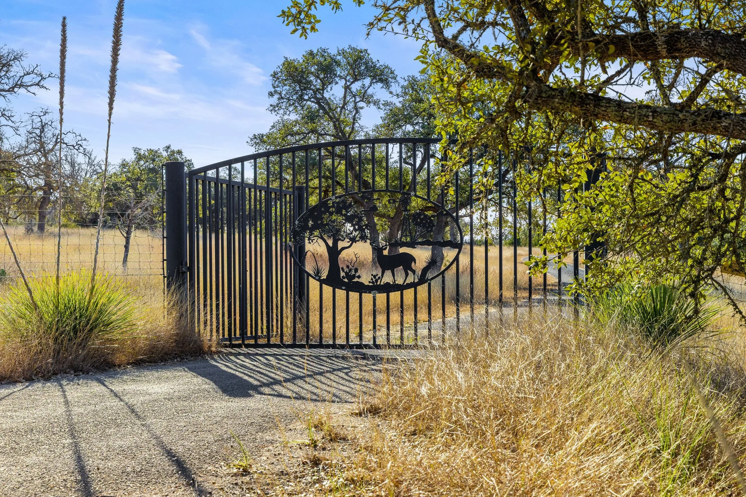A black metal gate with a circular design showing a silhouette of a deer, trees, and plants, situated in a rural area with grass, trees, and a partly cloudy sky.