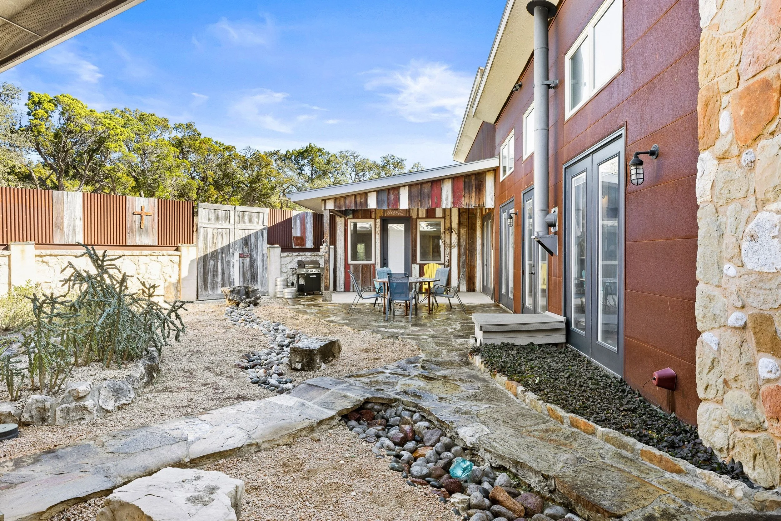 A backyard patio area with outdoor furniture, a stone pathway, a barbecue grill, and a fenced yard, with a partly cloudy blue sky overhead.