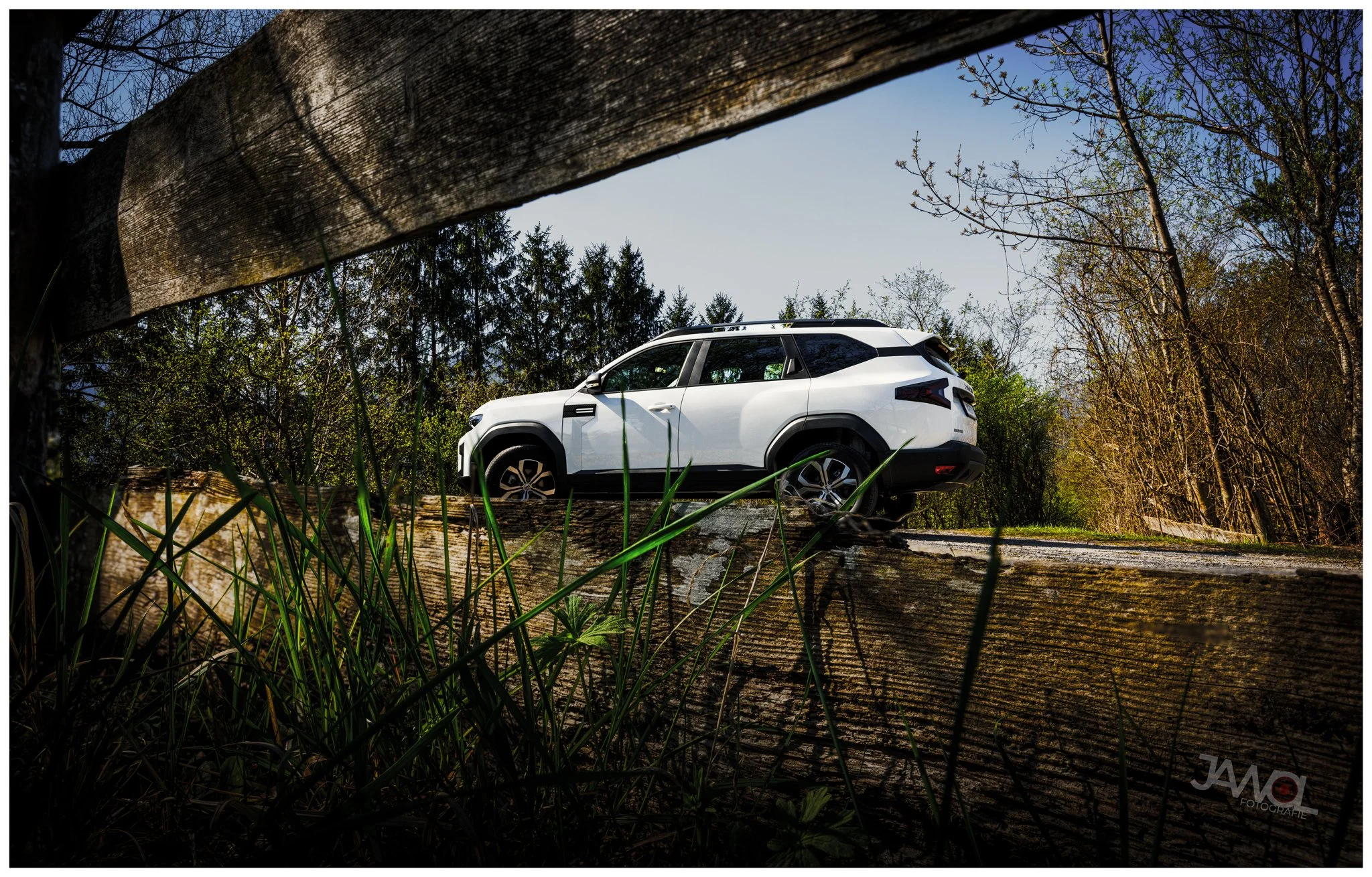 Ein weißes Auto steht auf einer Holzbrücke, umgeben von Bäumen und Gras, aufgenommen aus niedriger Perspektive durch eine Lücke im Holz.