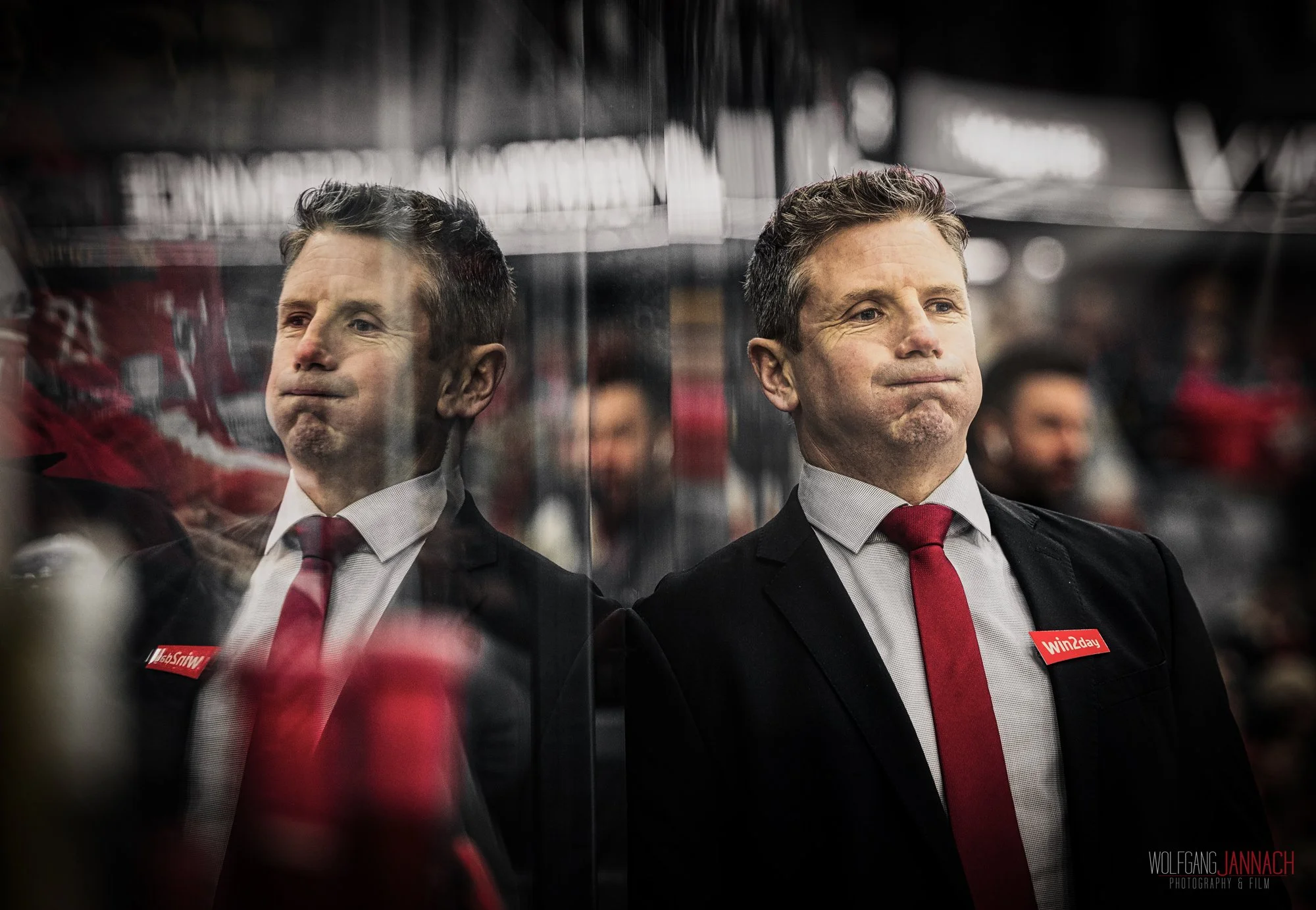 Ein Mann in formeller Kleidung mit schmaler Krawatte schaut nach oben, während er auf der anderen Seite eine spiegelnde Glasfläche betrachtet, die sein Spiegelbild zeigt. Es ist ein Innenraum mit Menschen im Hintergrund, die vermutlich bei einem Spor